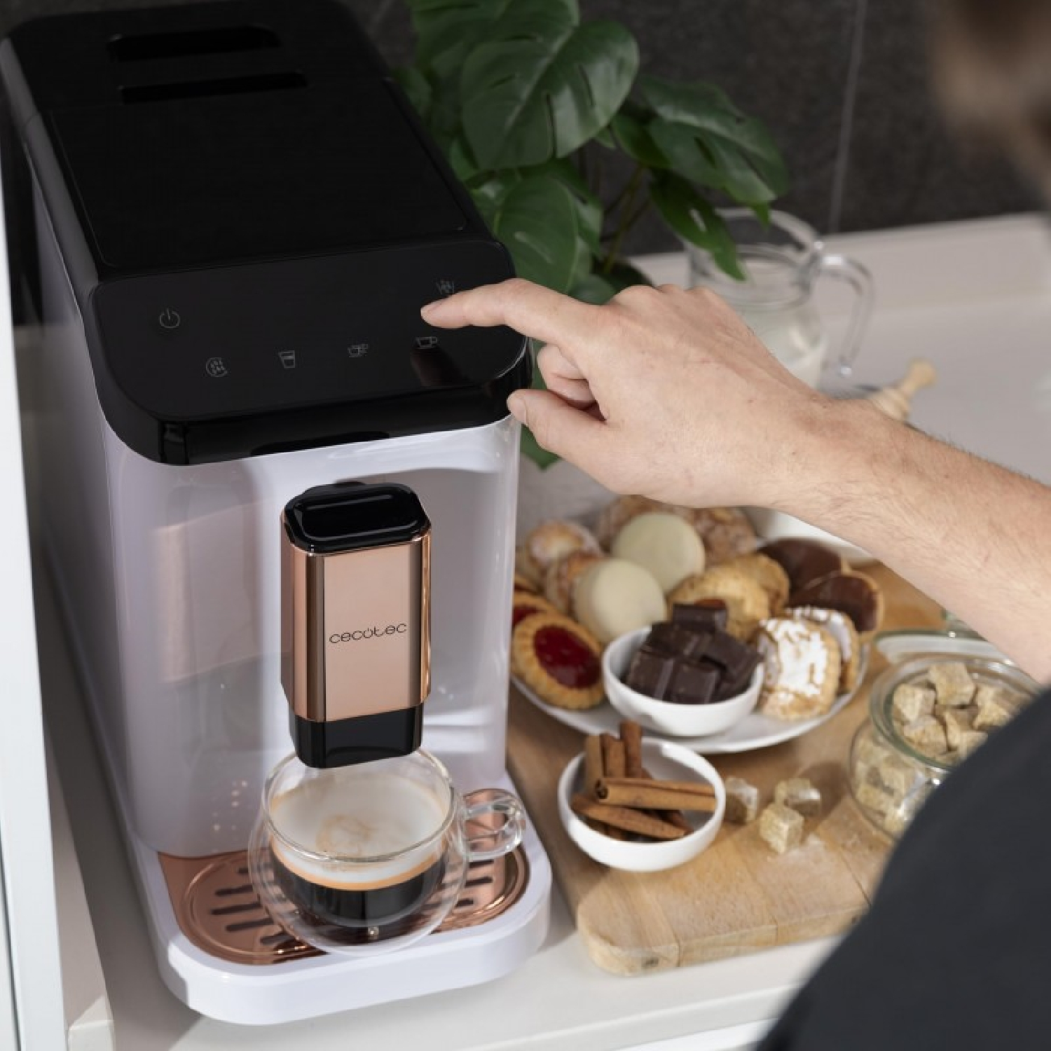 Cafetera dispensando café en un vaso, con aperitivos y una planta de fondo.