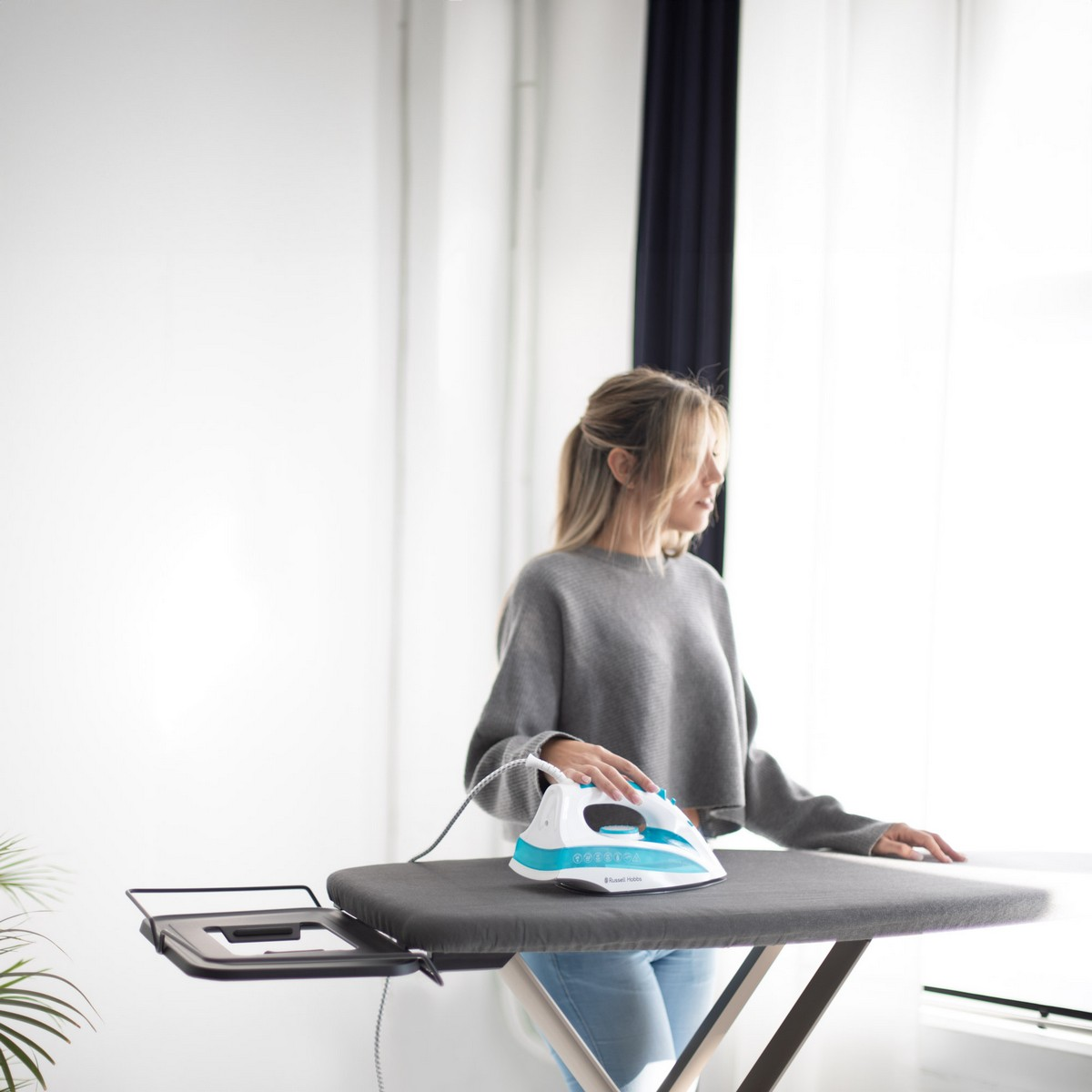 Mujer planchando ropa en una tabla de planchar cerca de una ventana. La plancha es azul claro y blanca.