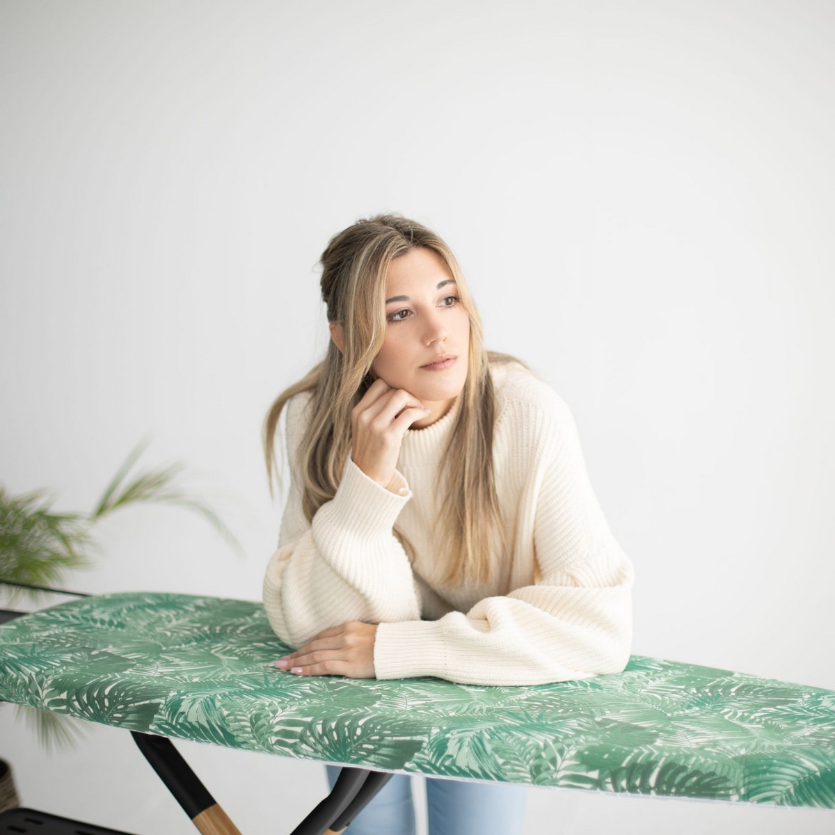 Mujer rubia apoyada en tabla de planchar, mirando hacia otro lado. Fondo blanco, estampado de hojas verdes.