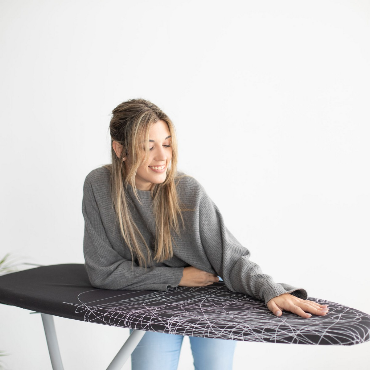 Mujer de pelo largo apoyada en una tabla de planchar, sonriendo. Fondo blanco.