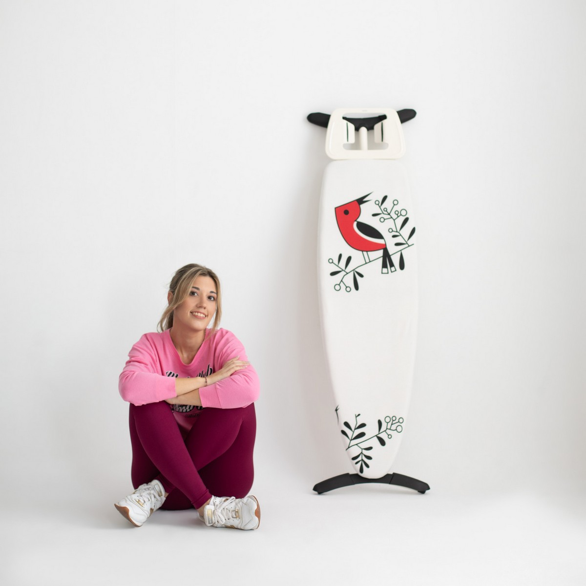 Una mujer sentada junto a una tabla de planchar blanca con diseño de pájaro sobre fondo blanco.