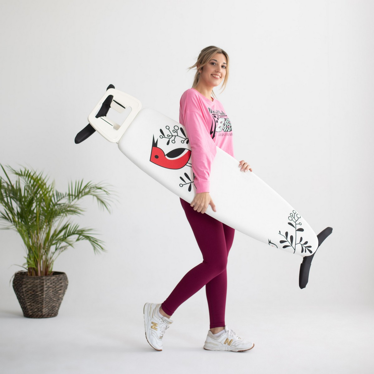 Una mujer cargando una tabla de planchar blanca con un diseño de pájaro rojo, sonriendo, sobre un fondo blanco.