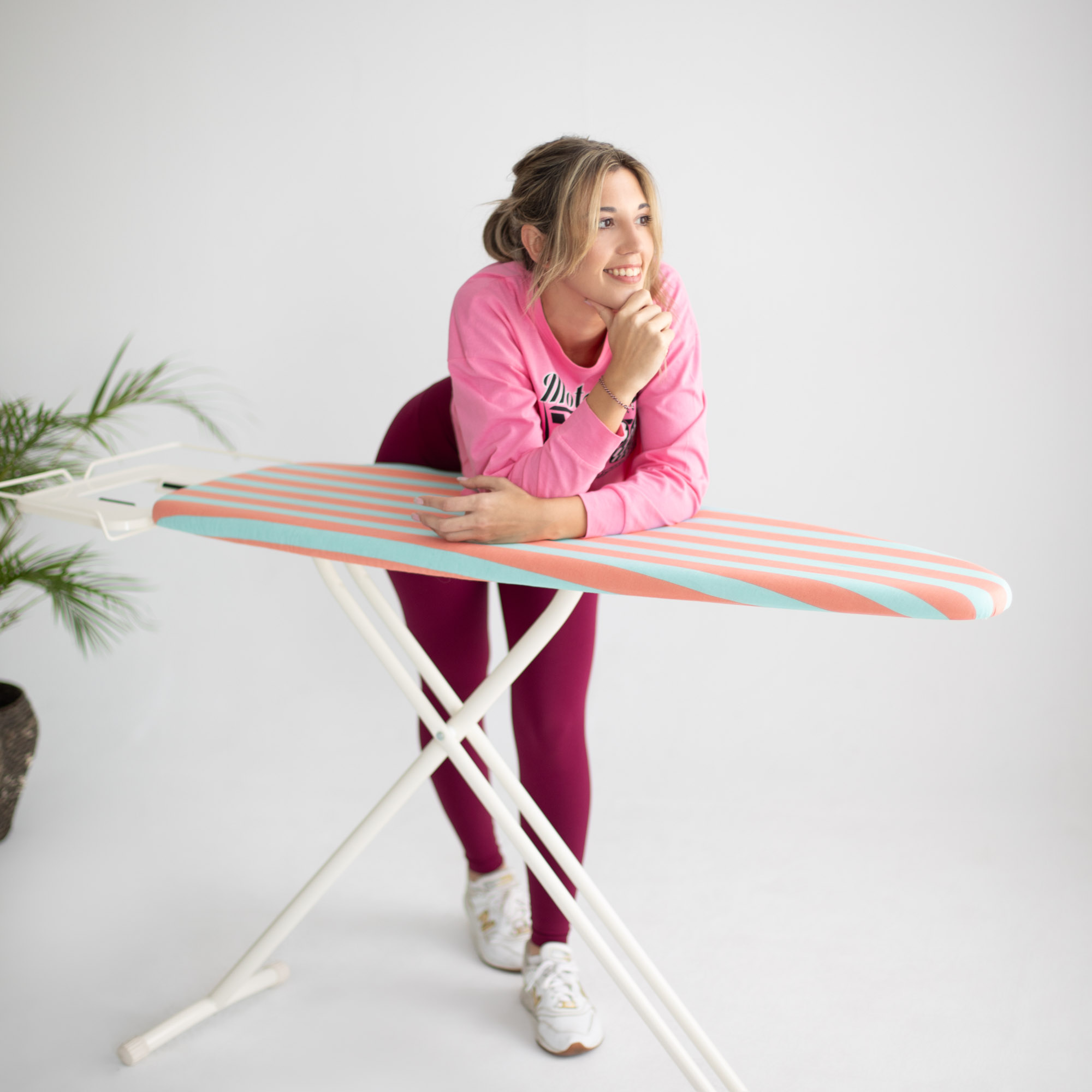 Una mujer apoyada en una tabla de planchar a rayas, sonriendo, con una planta en el fondo.
