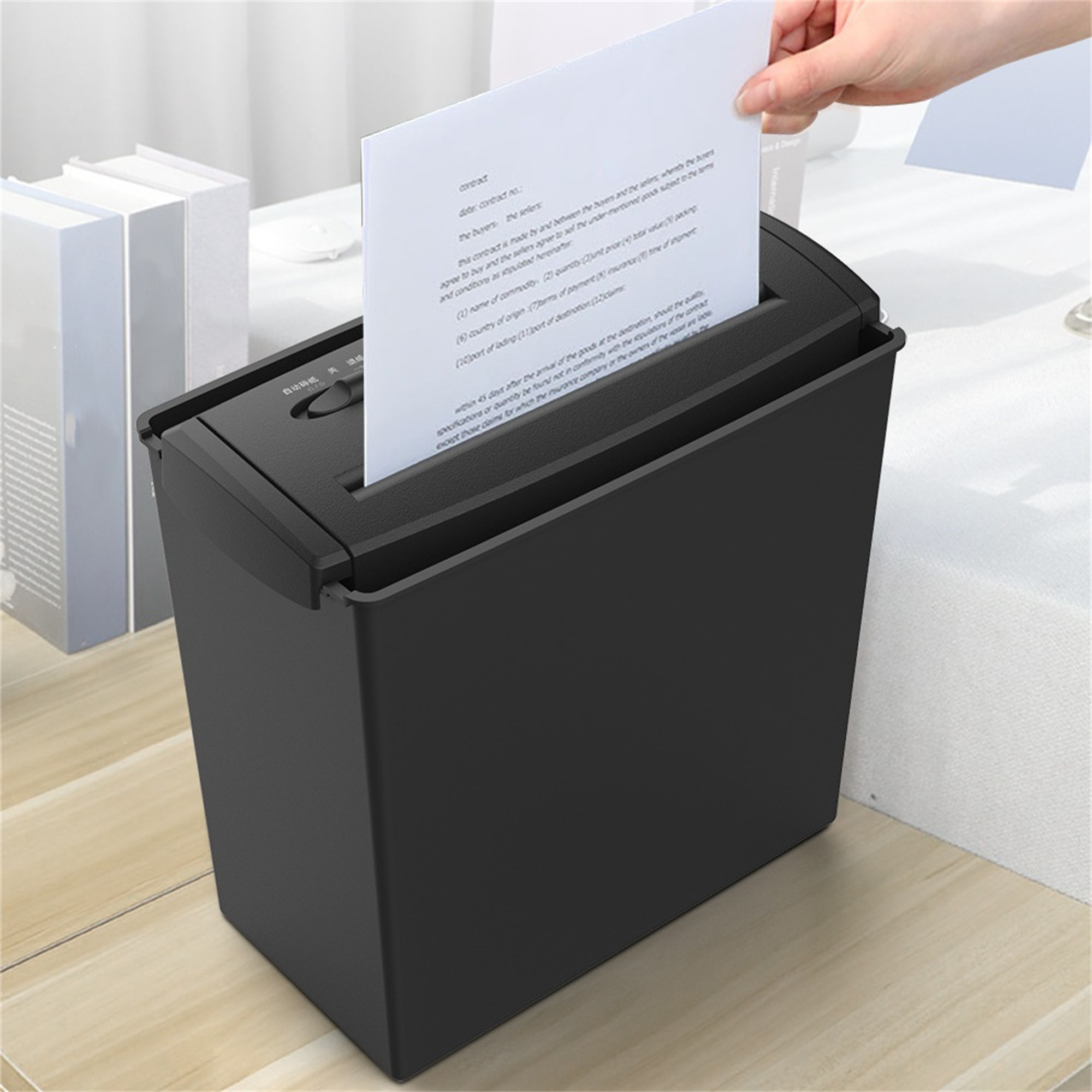 Schwarzer Aktenvernichter, in den eine Hand ein Dokument einlegt. Bücher im Hintergrund.