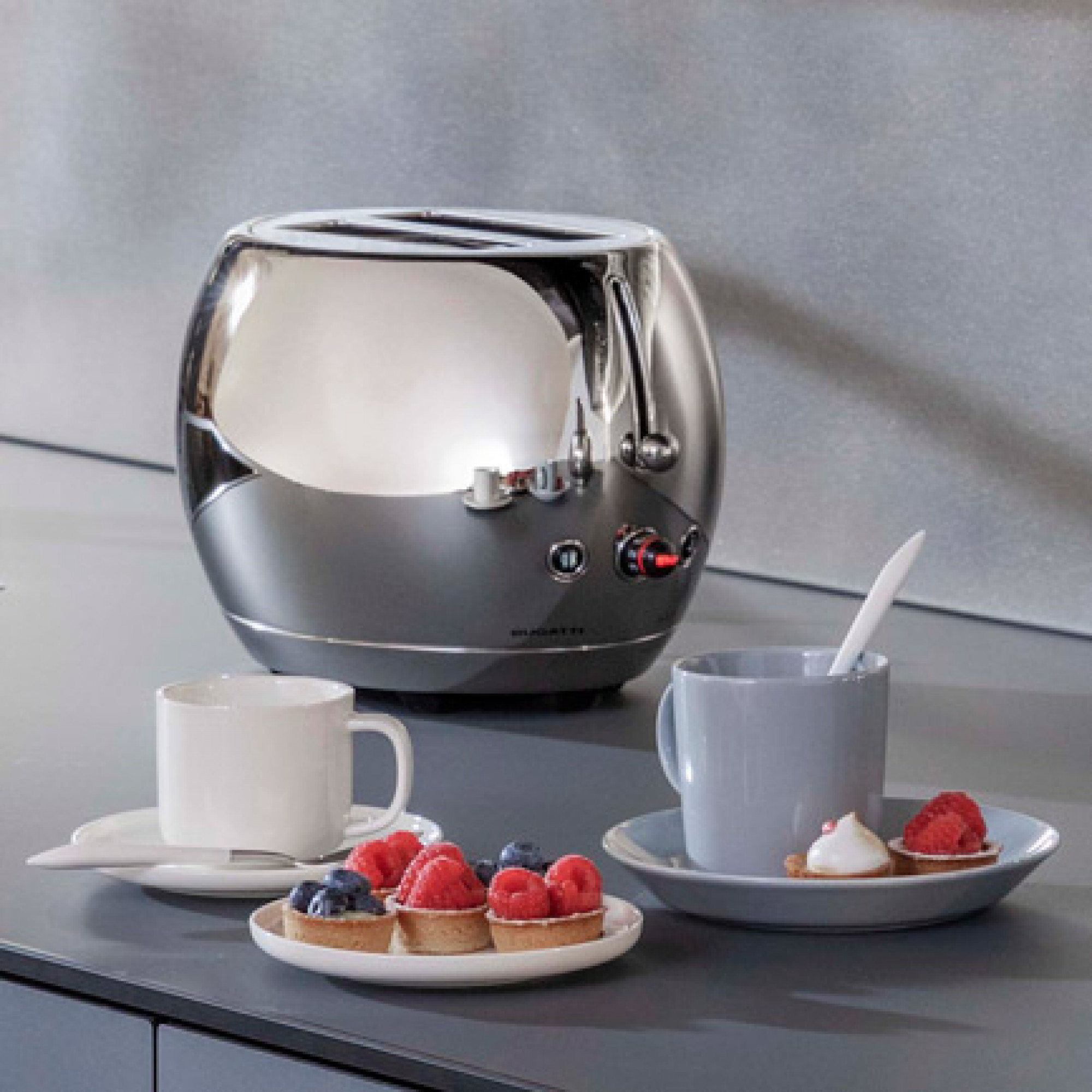 Tostapane argentato con luci rosse, tazze di caffè e pasticcini su un piano di lavoro grigio. Lo sfondo è un muro grigio.