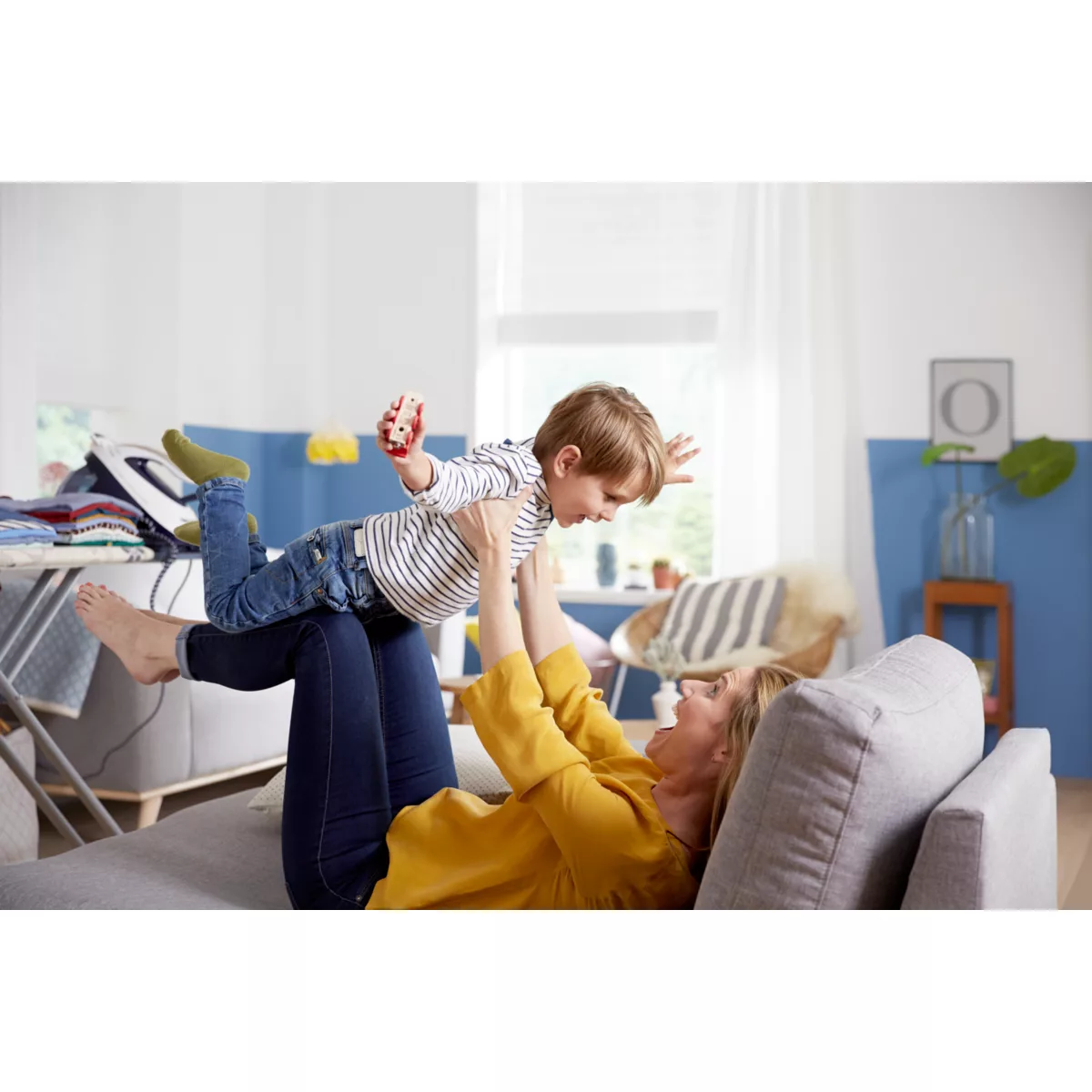 Eine Frau wirft ein Kind in die Luft, während sie auf einer Couch liegt. Kind hält Spielzeug.