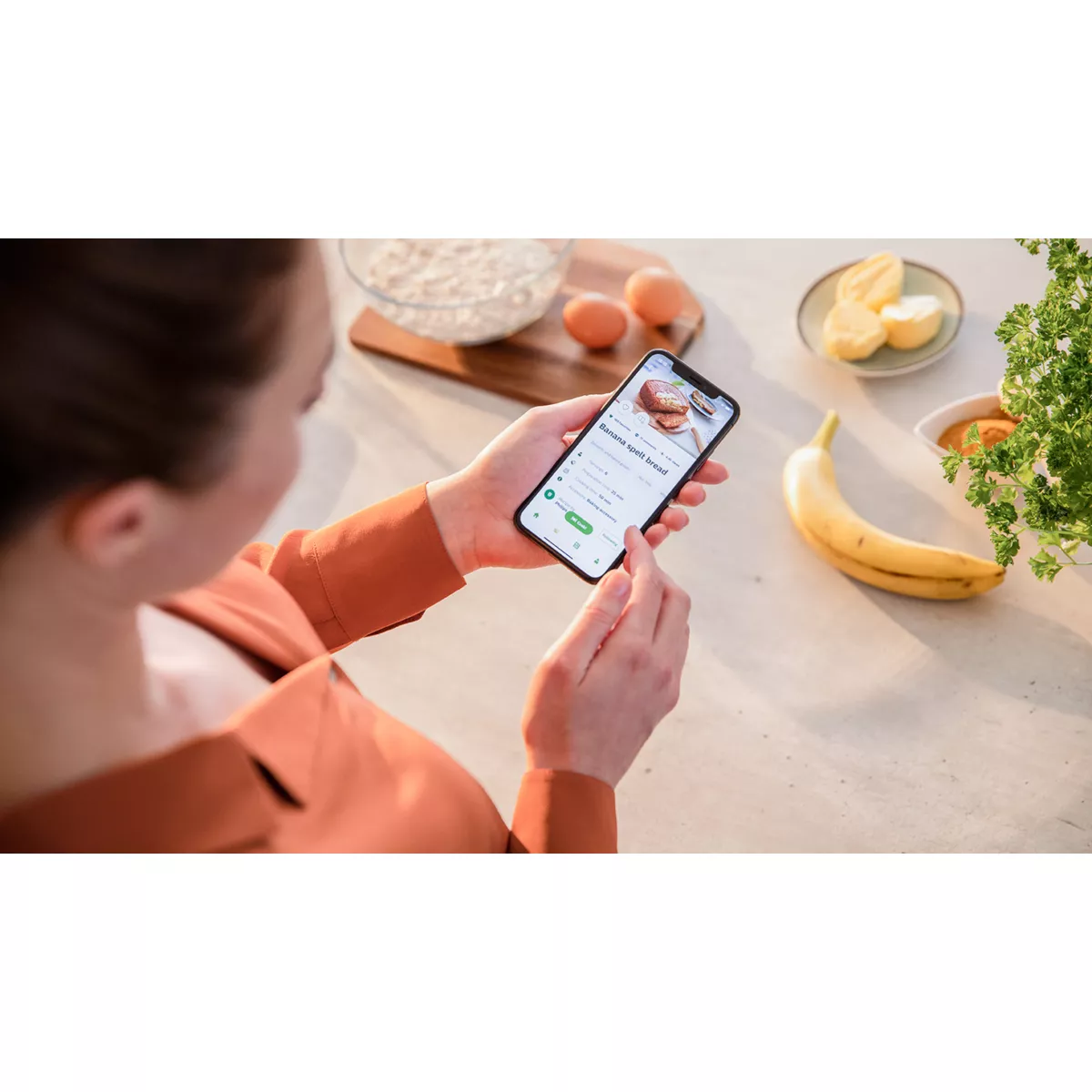 Frau hält ein Handy mit Rezept. Zutaten: Eier, Mehl, eine Banane und Zitronenscheiben.