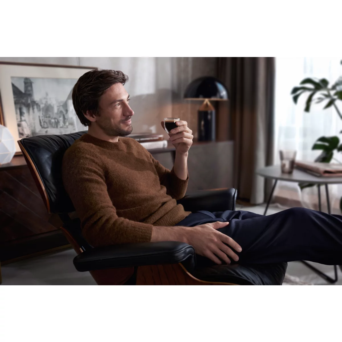 Mann sitzt, trinkt Kaffee, in modernem Wohnzimmer. Brauner Pullover, dunkle Hose.