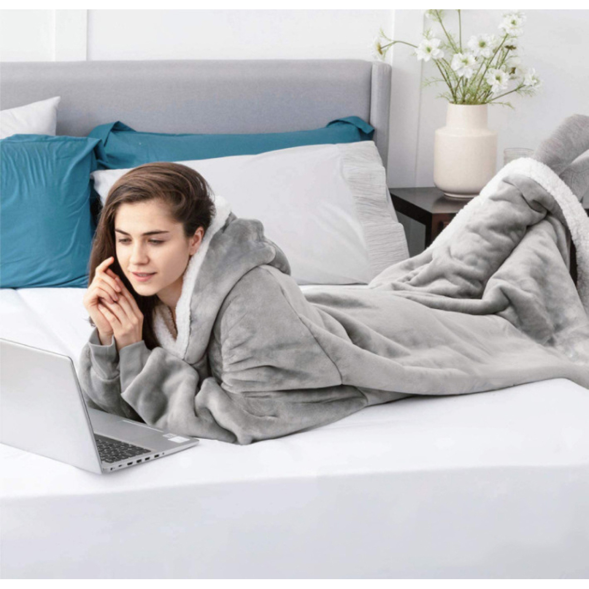 Frau in grauer Kapuzendecke benutzt Laptop im Bett. Vase mit Blumen im Hintergrund.