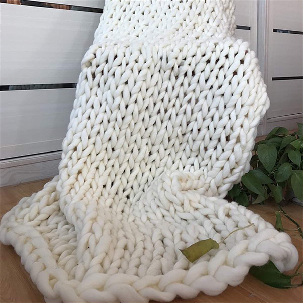 Eine grob gestrickte weiße Decke, die über eine Oberfläche drapiert ist, mit einer Pflanze im Hintergrund.