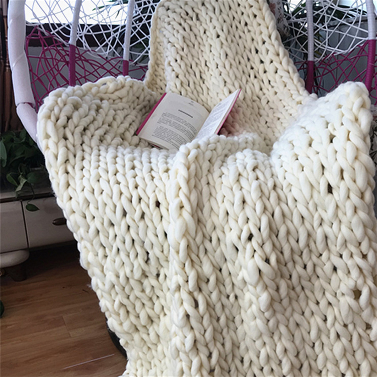 Eine cremefarbene, grob gestrickte Decke, die über einen Stuhl drapiert ist, mit einem Buch darauf.