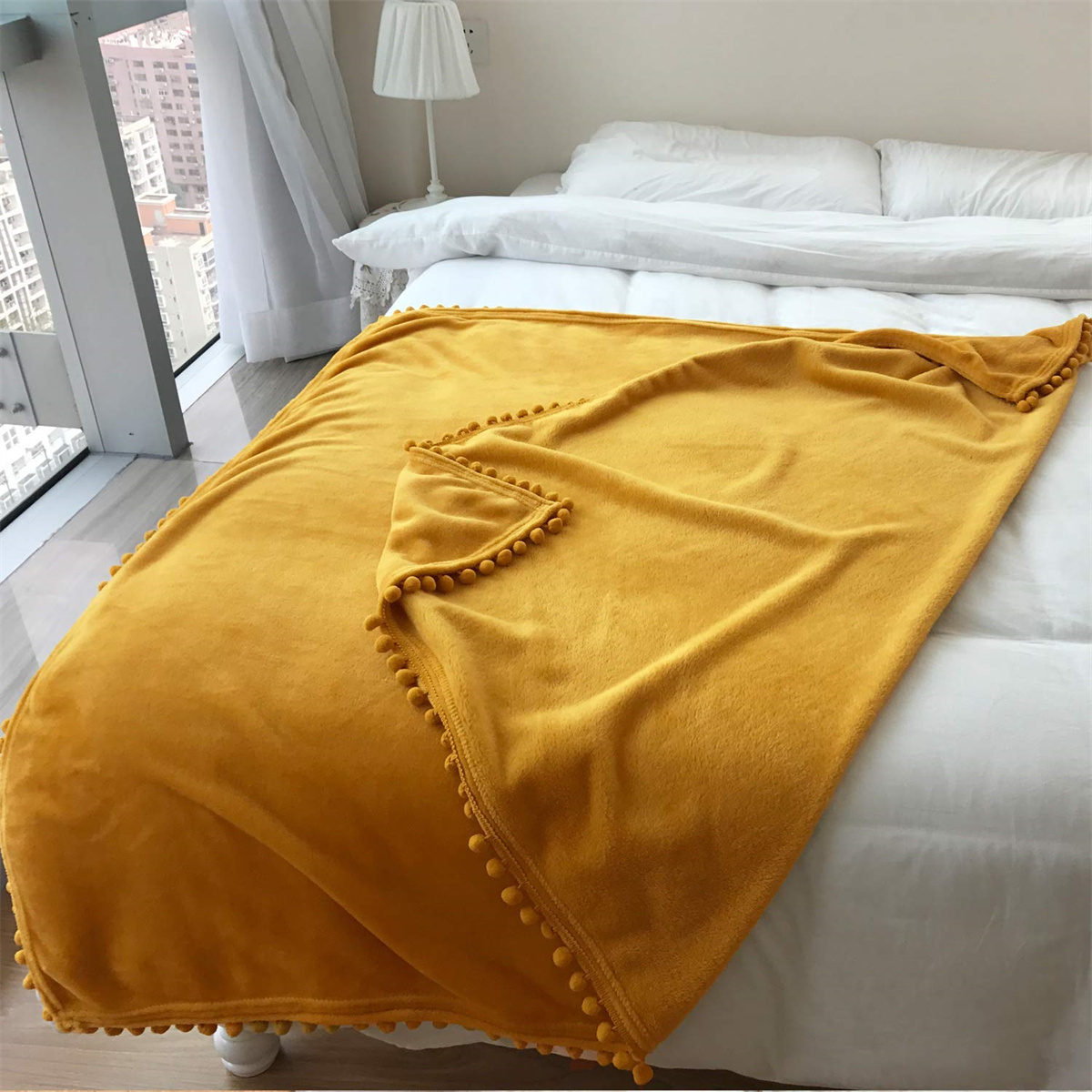 Eine gelbe Decke mit Bommeln liegt auf einem weißen Bett. Im Hintergrund ist ein Fenster zu sehen.