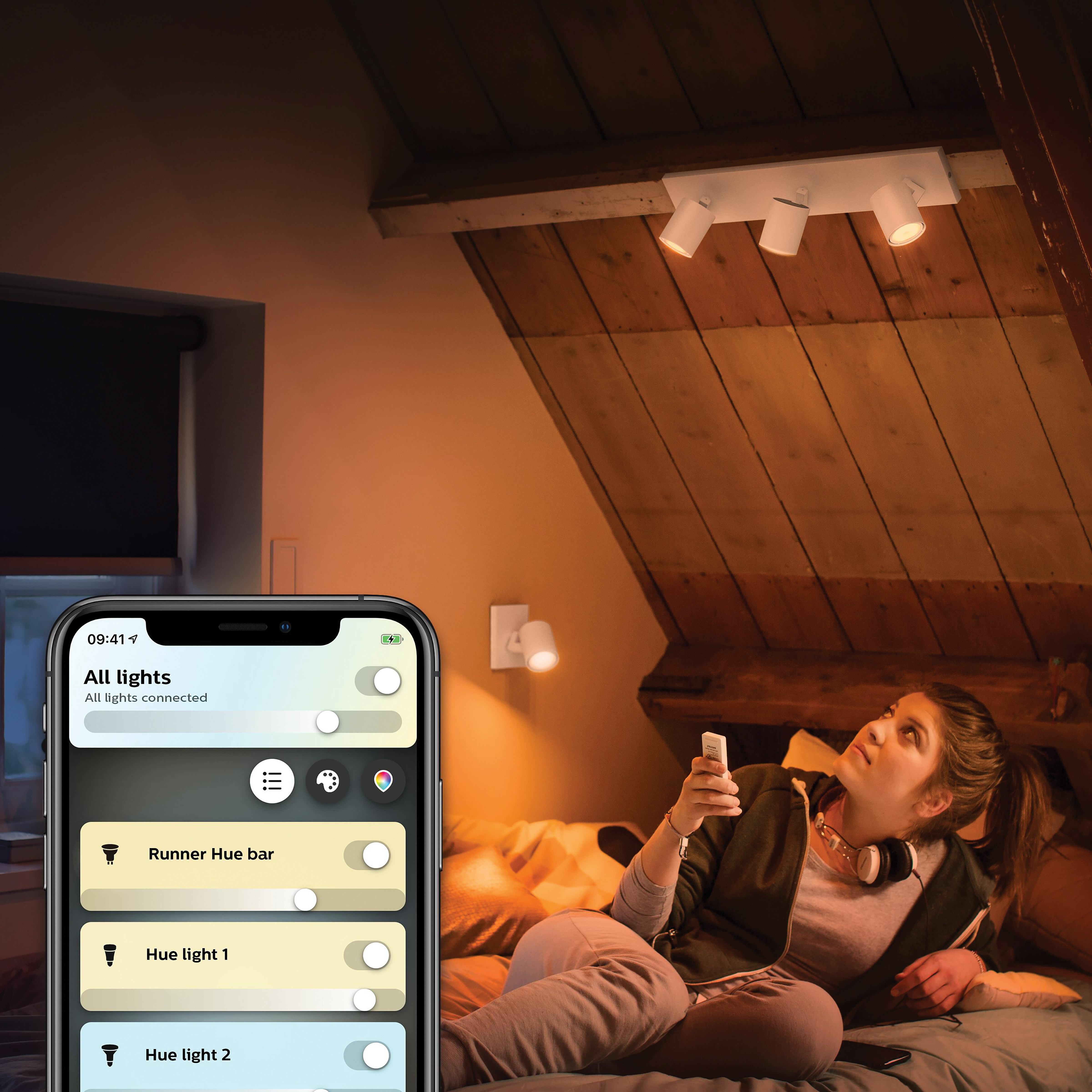 Eine Frau blickt auf drei Strahler an einer Holzdecke, hält ein Telefon mit einer App.
