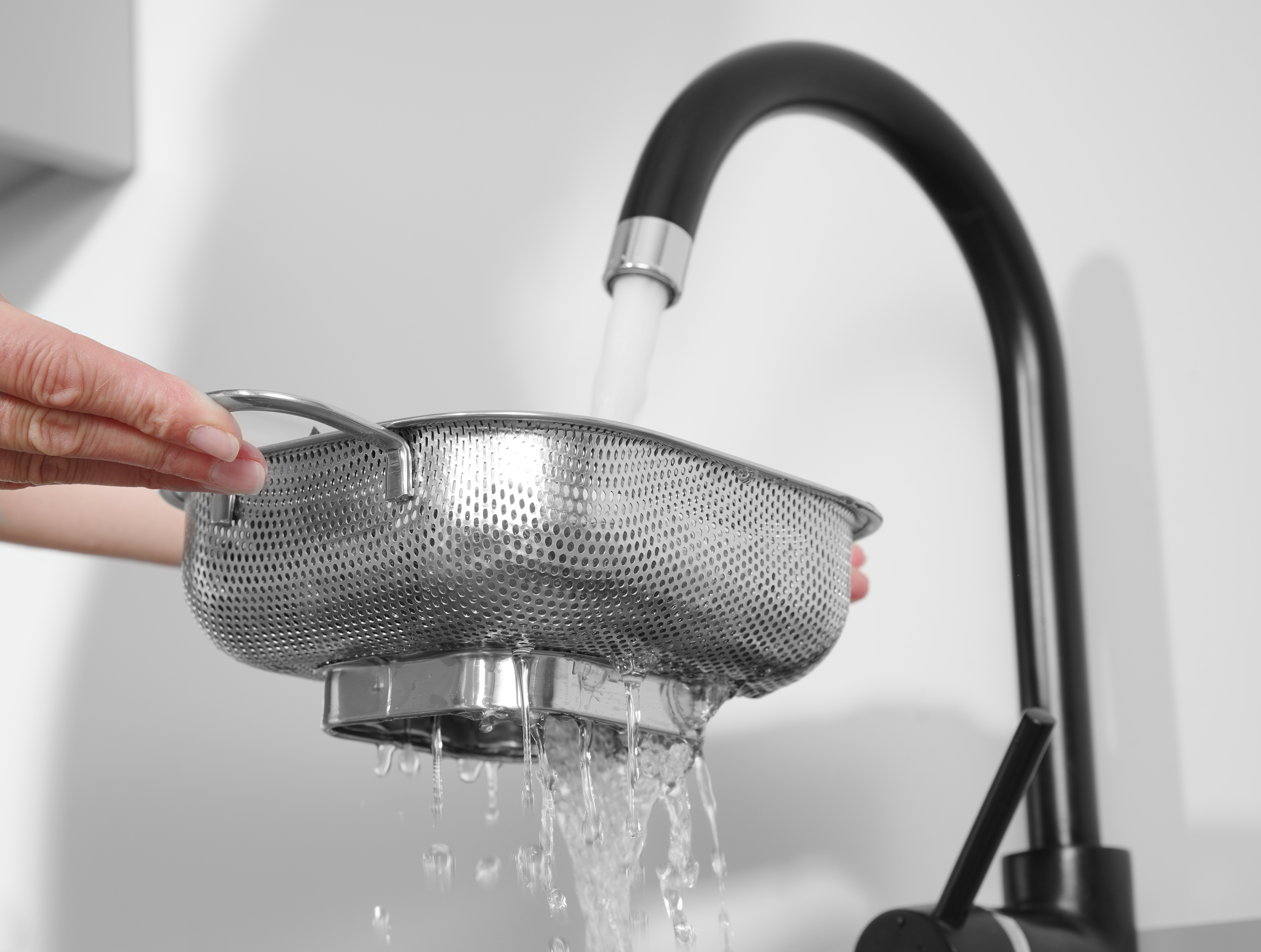 Un colino viene tenuto sotto un rubinetto con acqua che scorre attraverso. Il colino è in acciaio, il rubinetto è nero.