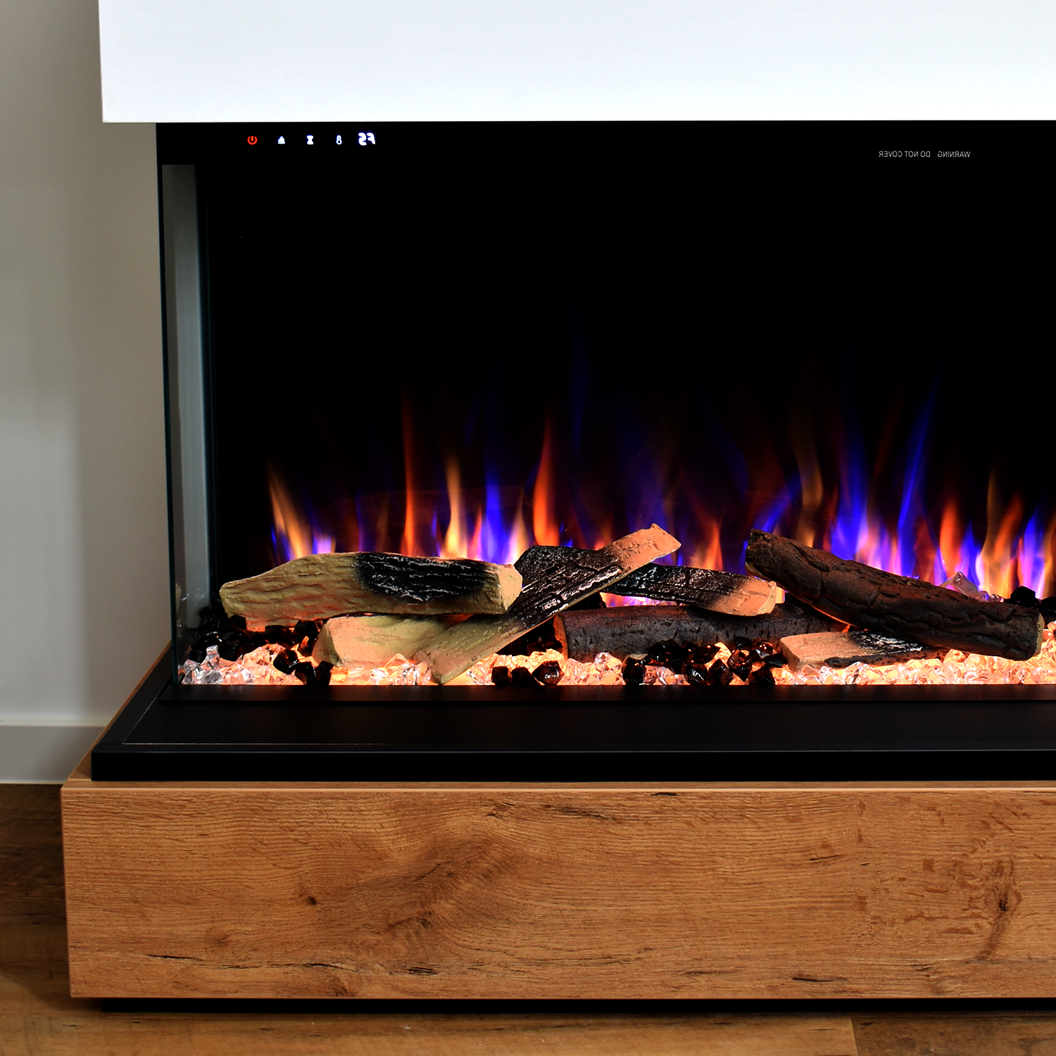 Elektrischer Kamin mit Holzscheiten und blau/orangenen Flammen. Holzsockel, schwarzer Rahmen, weißer Deckel.