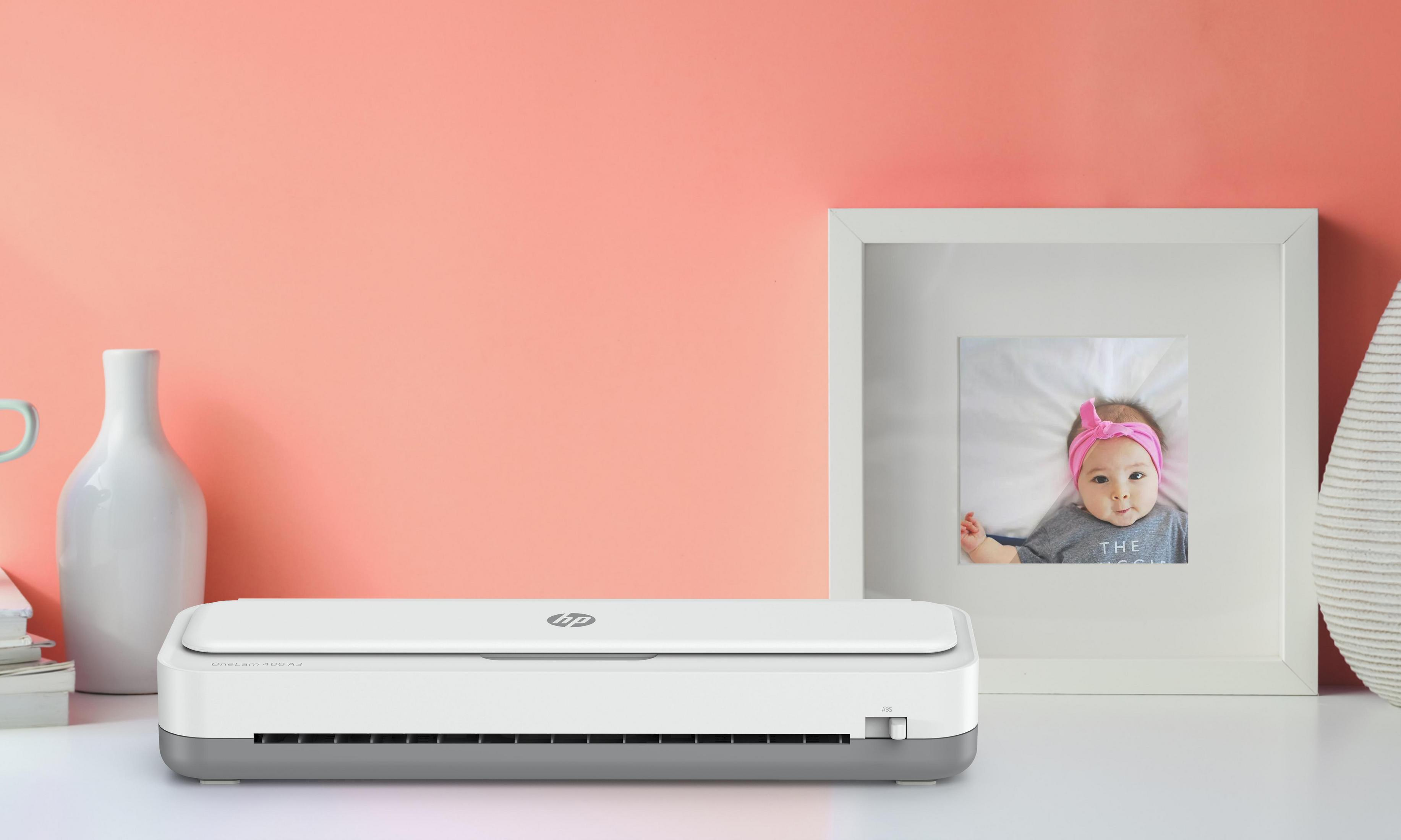 Biała drukarka stoi na białym stole z oprawionym zdjęciem dziecka i białym wazonem w tle.