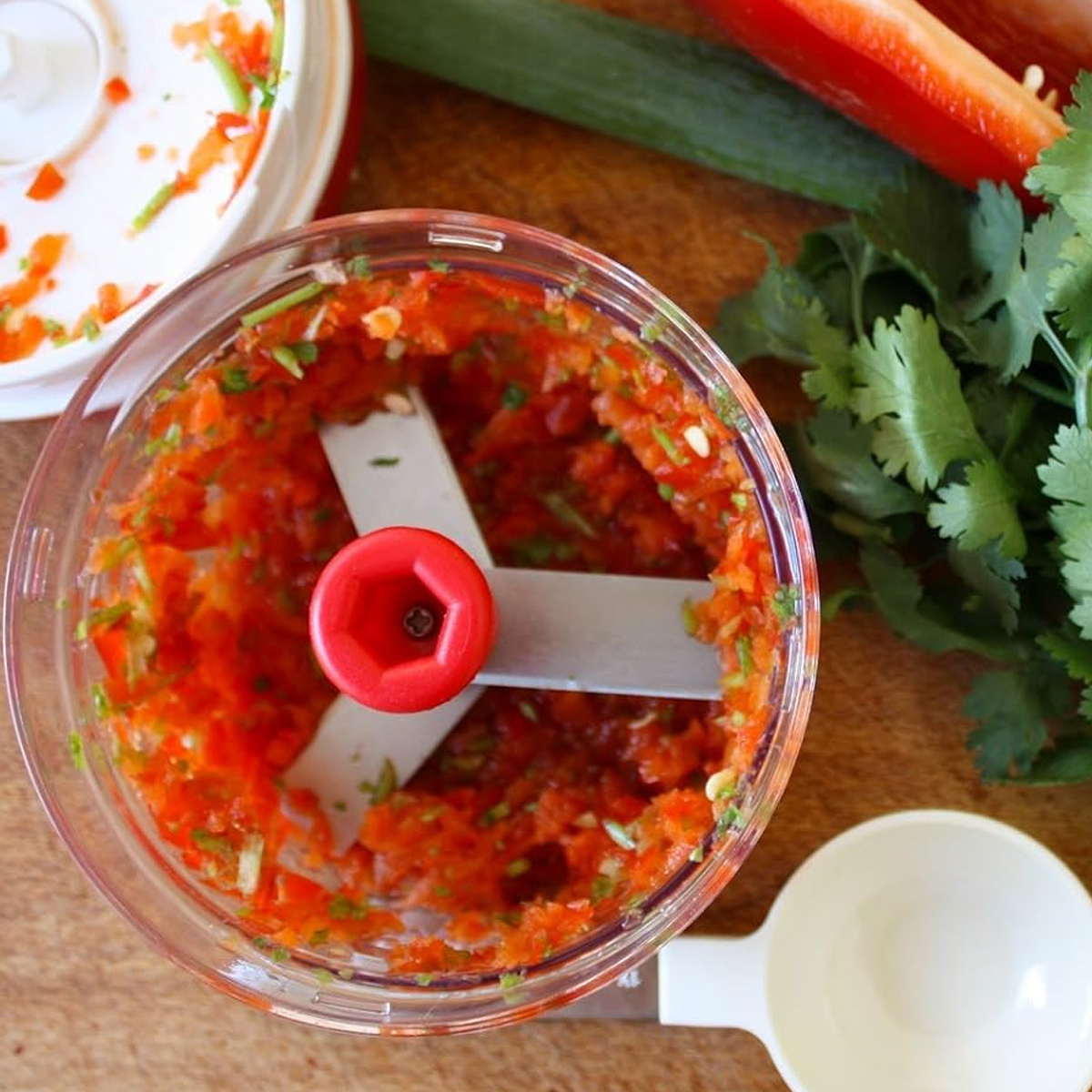 Rote Paprika- und Koriander-Mischung in einem transparenten Foodprocessor, mit Löffel und Schneidebrett.