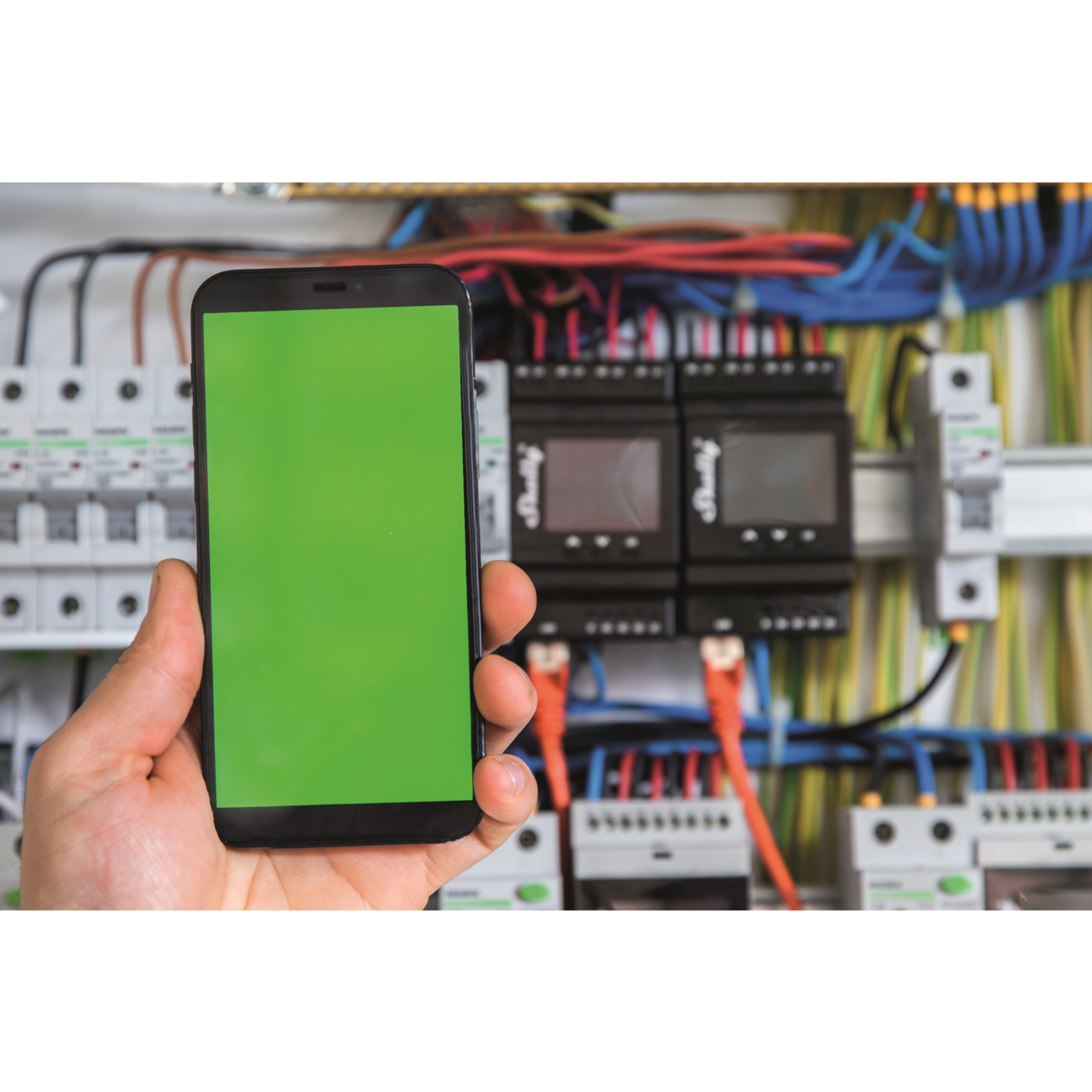 Ręka trzymająca telefon z zielonym ekranem przed komponentami elektrycznymi i przewodami.