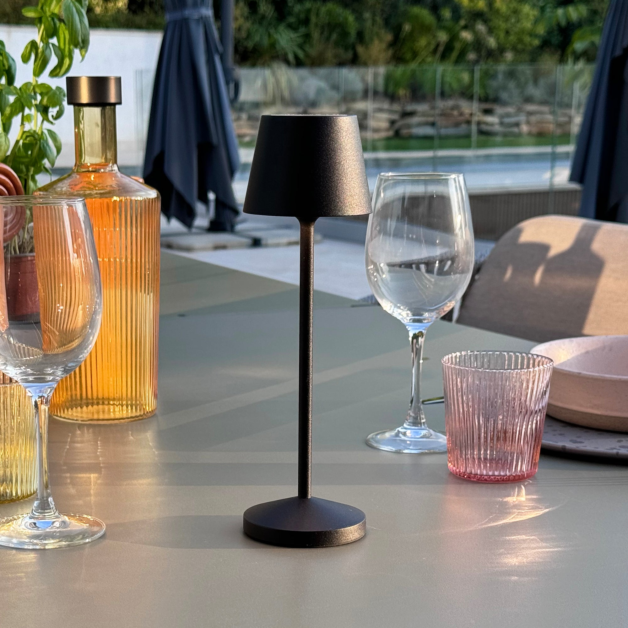 Eine schwarze Lampe mit schwarzem Lampenschirm auf einem Tisch mit Gläsern und Flaschen.