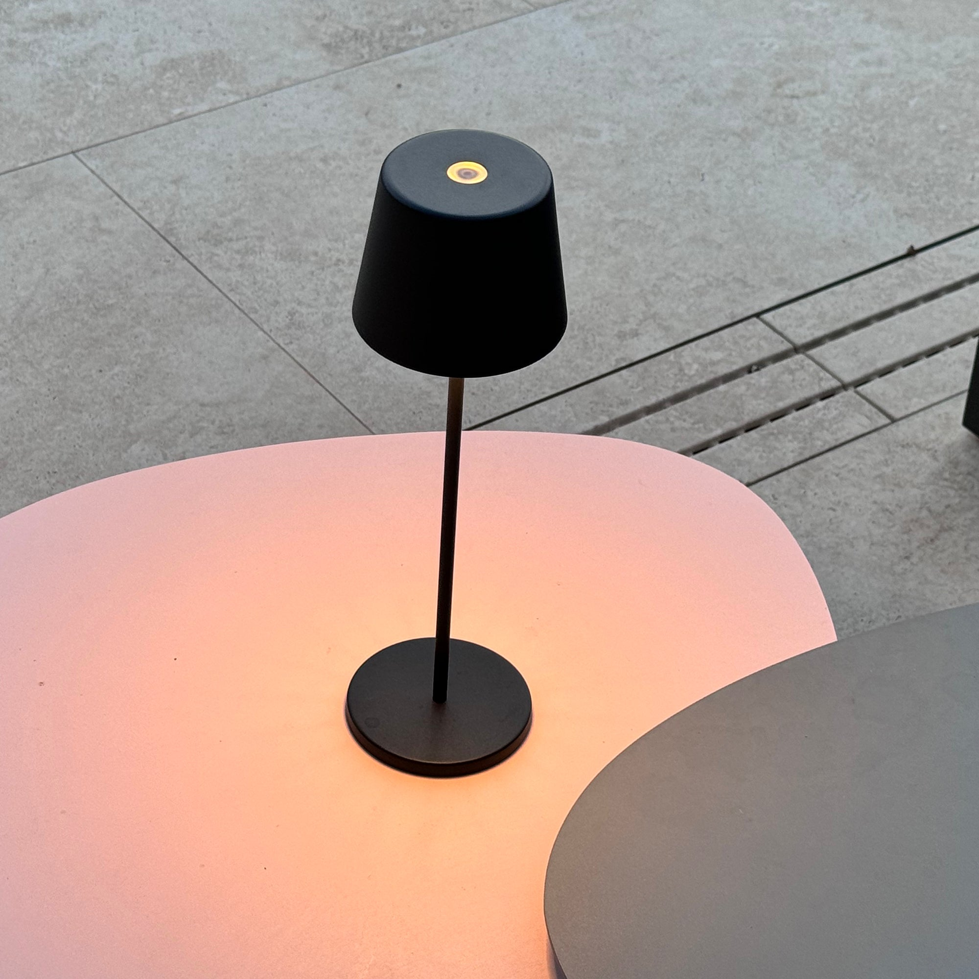 Schwarze Lampe mit rundem Fuß, und einem rosa Tisch. Hintergrund ist eine graue Oberfläche.