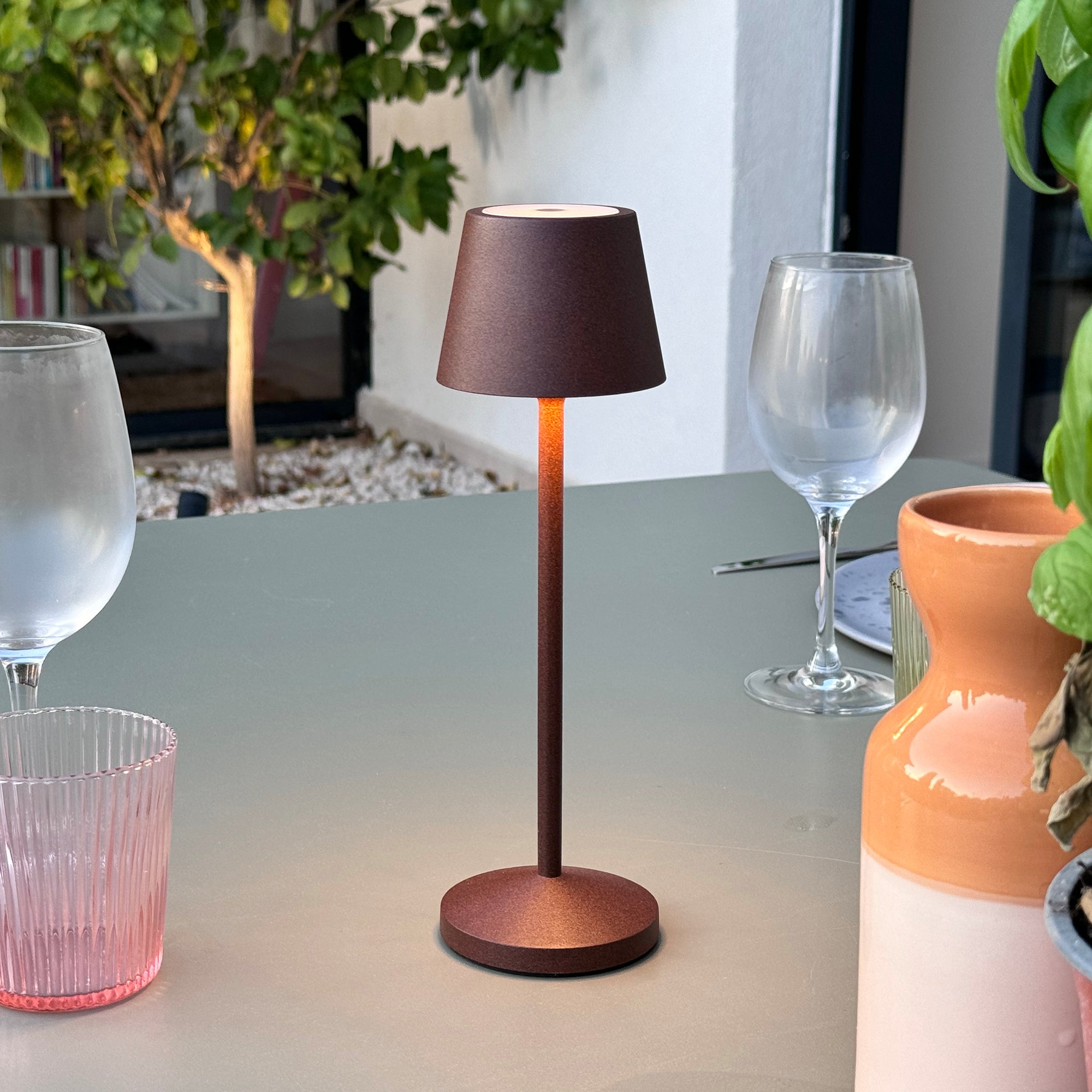 Eine kleine braune Tischlampe auf einem Tisch mit Weinglas und dekorativem Topf.