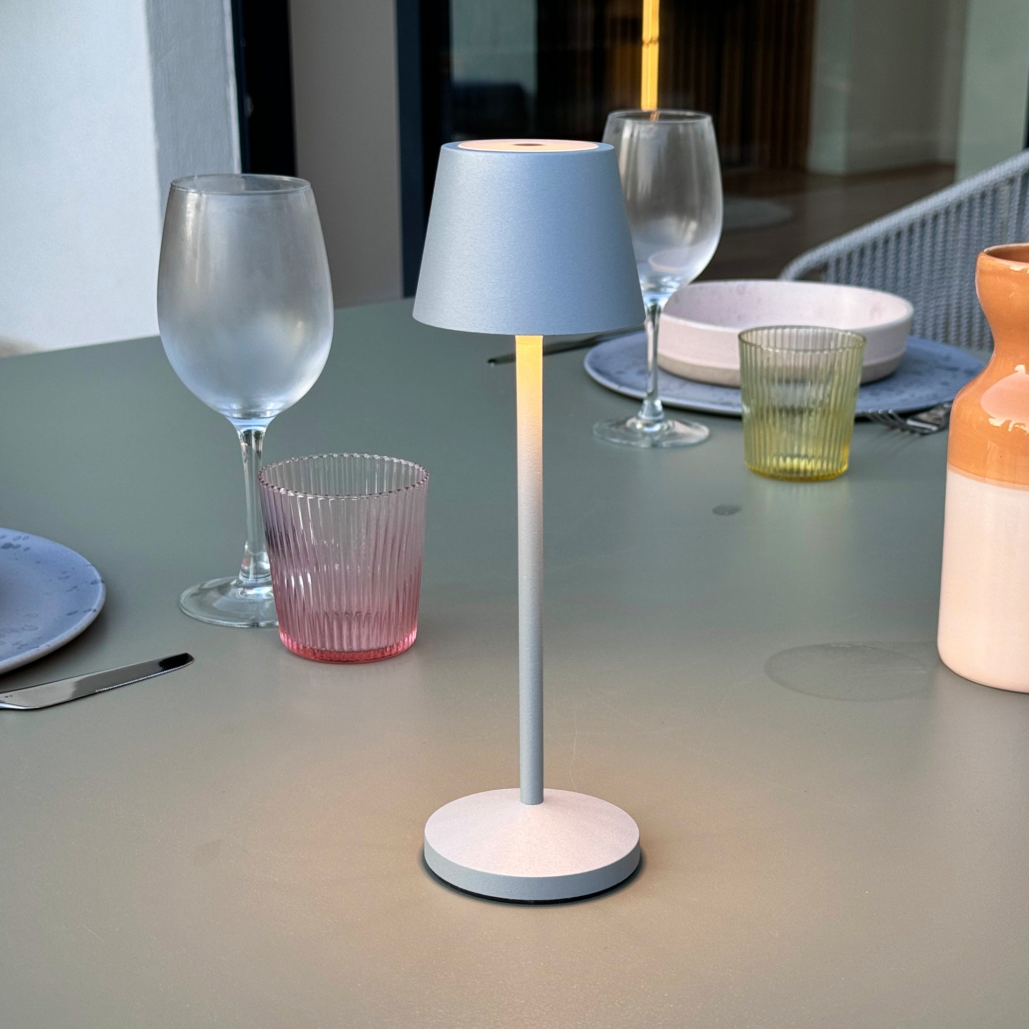 Ein grauer Tisch mit einer Lampe, Gläsern und einer Vase. Die Lampe hat einen grauen Stiel und einen hellblauen Schirm.