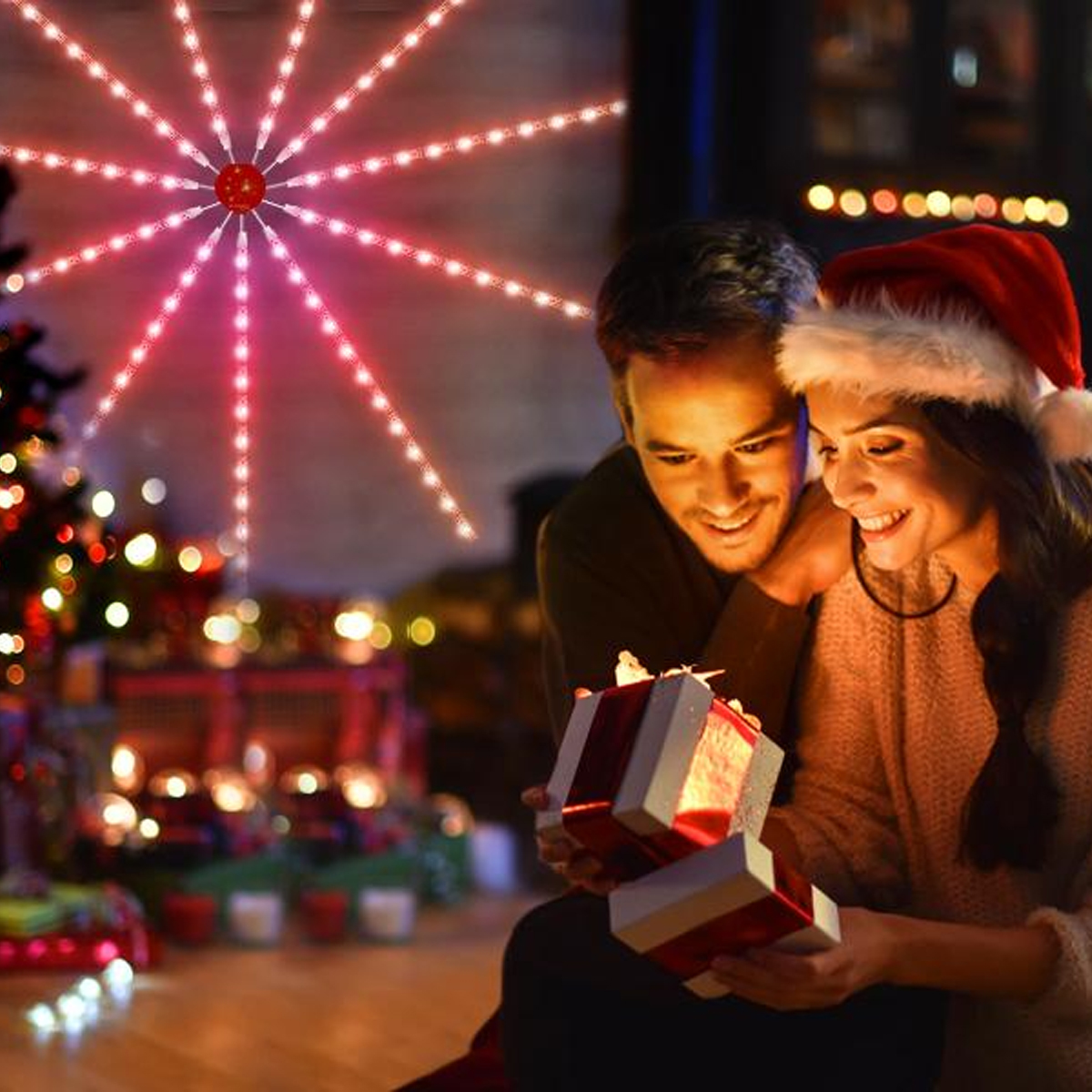 Paar öffnet Geschenke unter Weihnachtsbeleuchtung. Über ihnen befindet sich eine rote sternförmige Leuchte.