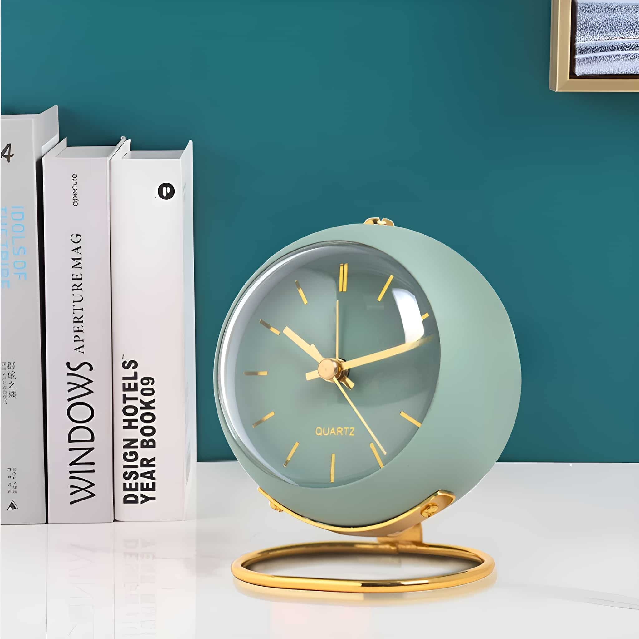 Grüne Uhr mit goldenem Sockel und goldenen Zeigern, weiße Bücher und eine türkisfarbene Wand im Hintergrund.