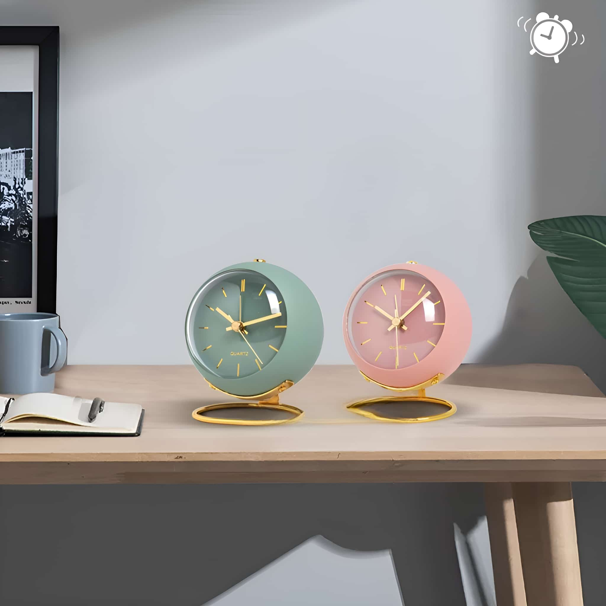 Zwei Uhren im Retro-Stil mit goldenen Ständern, eine mintgrün, eine rosa, auf einem Holztisch.