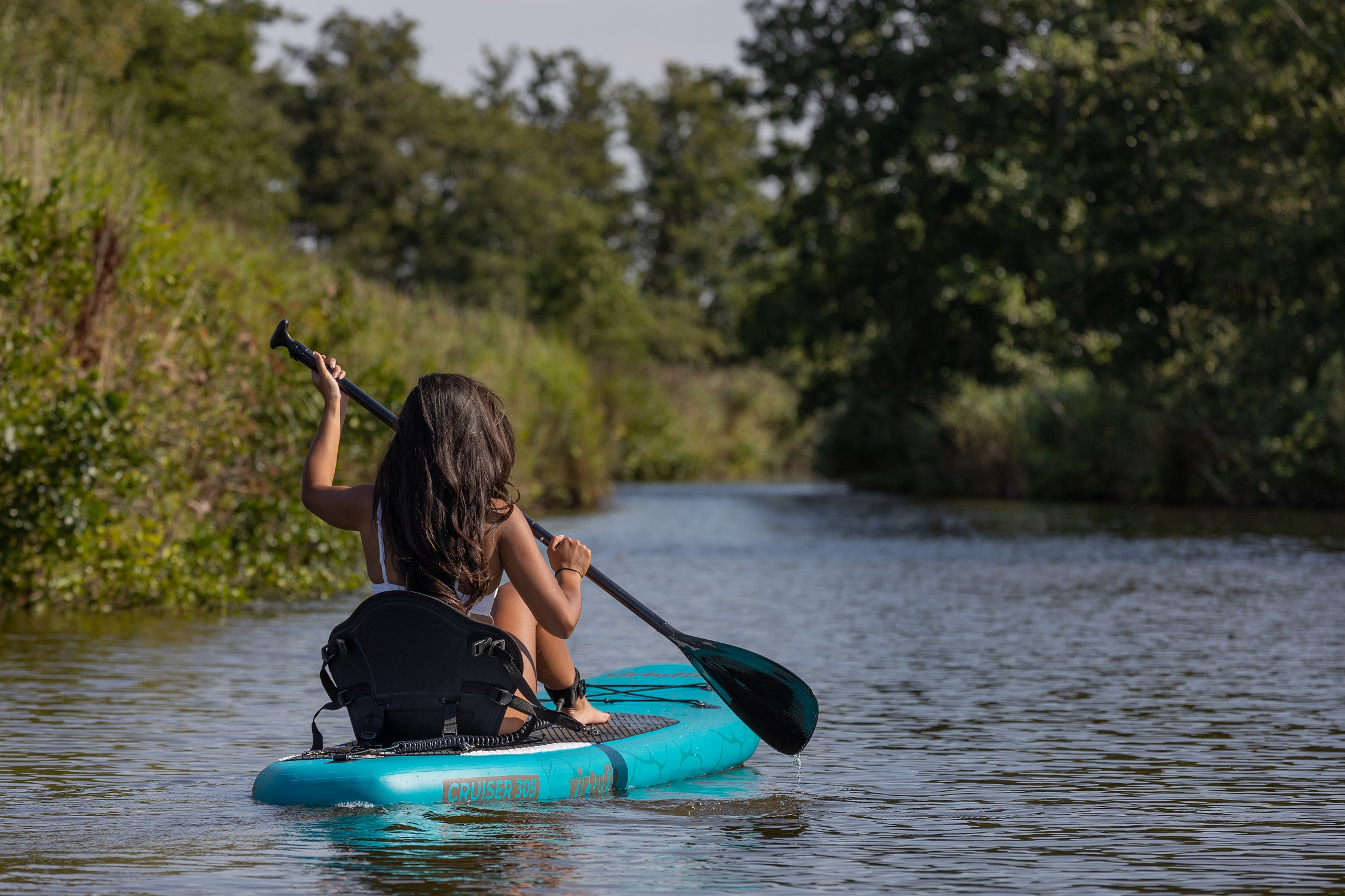 Frau auf einem Paddleboard, paddelt auf einem Fluss. Bäume säumen die Ufer.