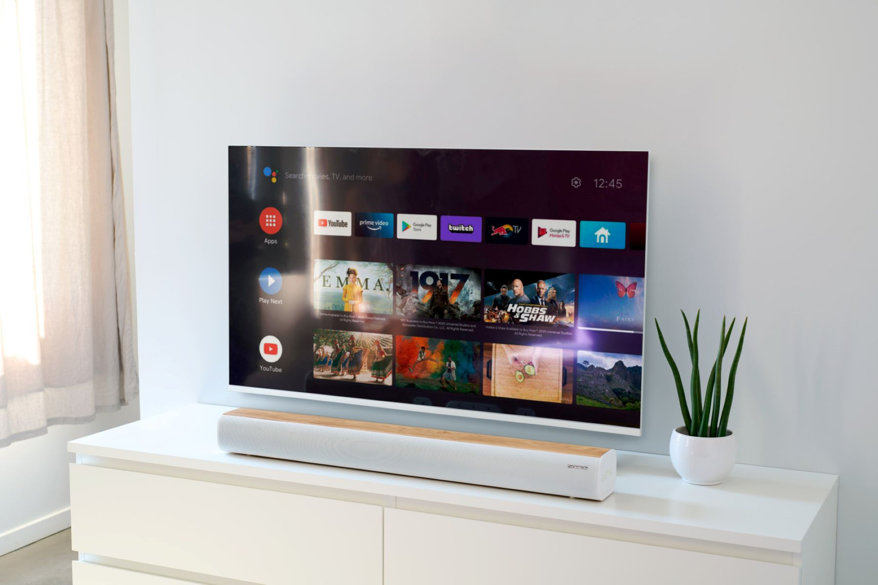 Ekran telewizora wyświetlający interfejs Smart TV i soundbar na białej szufladzie. Roślina po prawej.