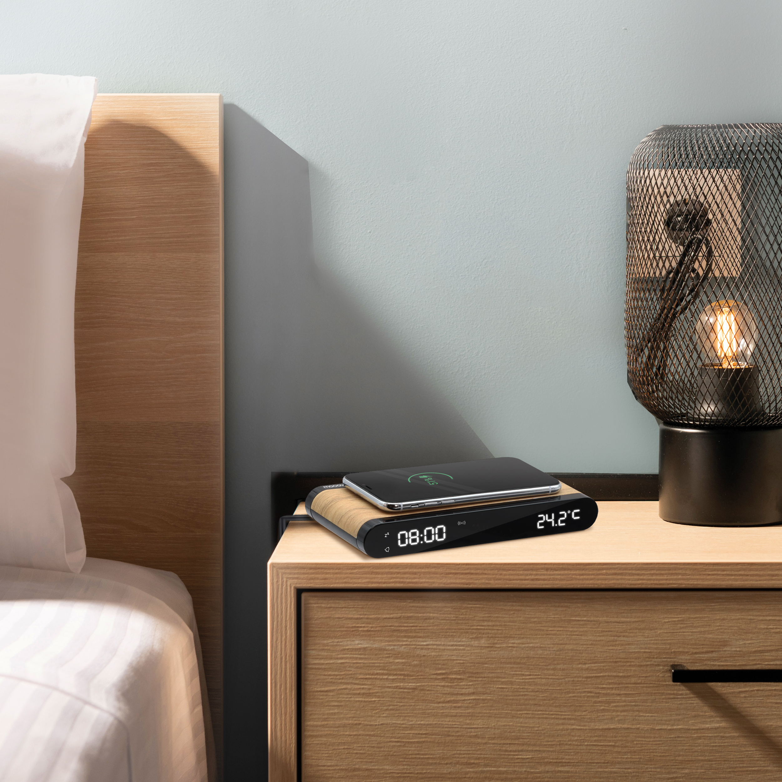 Un teléfono se carga en una mesita de noche de madera. Una lámpara está al lado, en una mesita de noche de madera.
