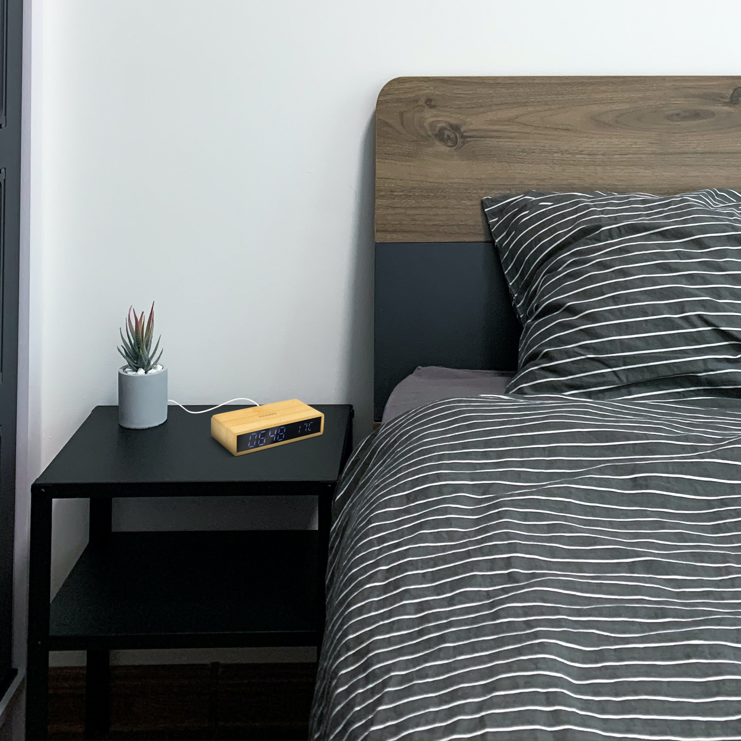 Mesita de noche de madera con reloj digital, planta y libros. Ropa de cama a rayas y cabecero de madera.
