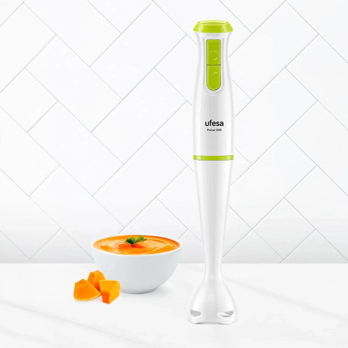 Batidora de mano Ufesa con cuerpo blanco y detalles verdes. Un cuenco de sopa naranja y calabaza en cubos.