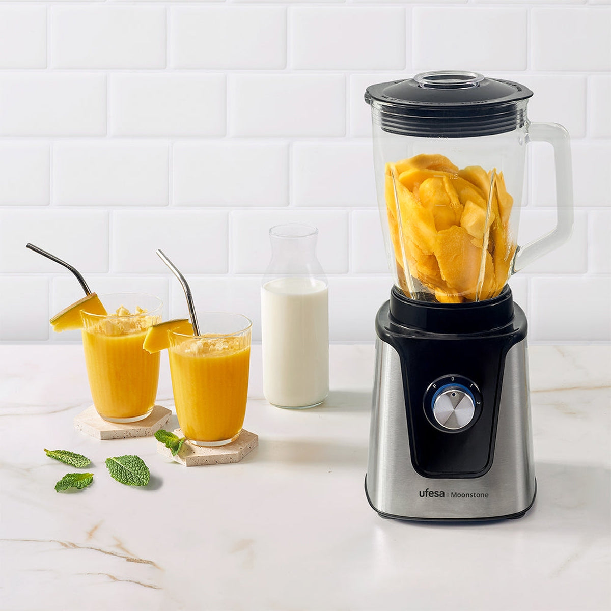 Licuadora con trozos de mango, dos batidos de mango y una botella de leche. Fondo de azulejos blancos.