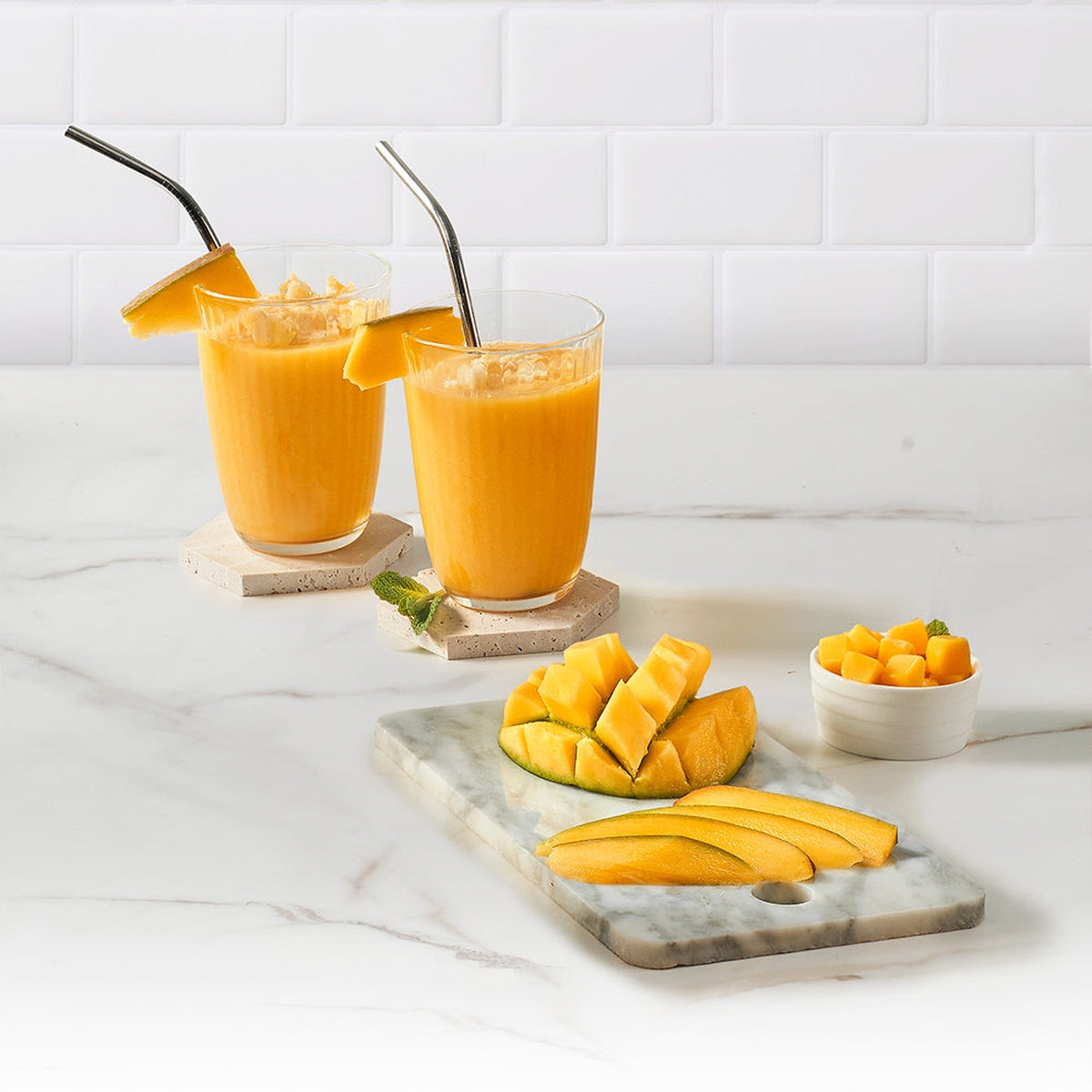 Dos batidos de mango con pajitas, rodajas de mango en una tabla de mármol y cubos de mango.