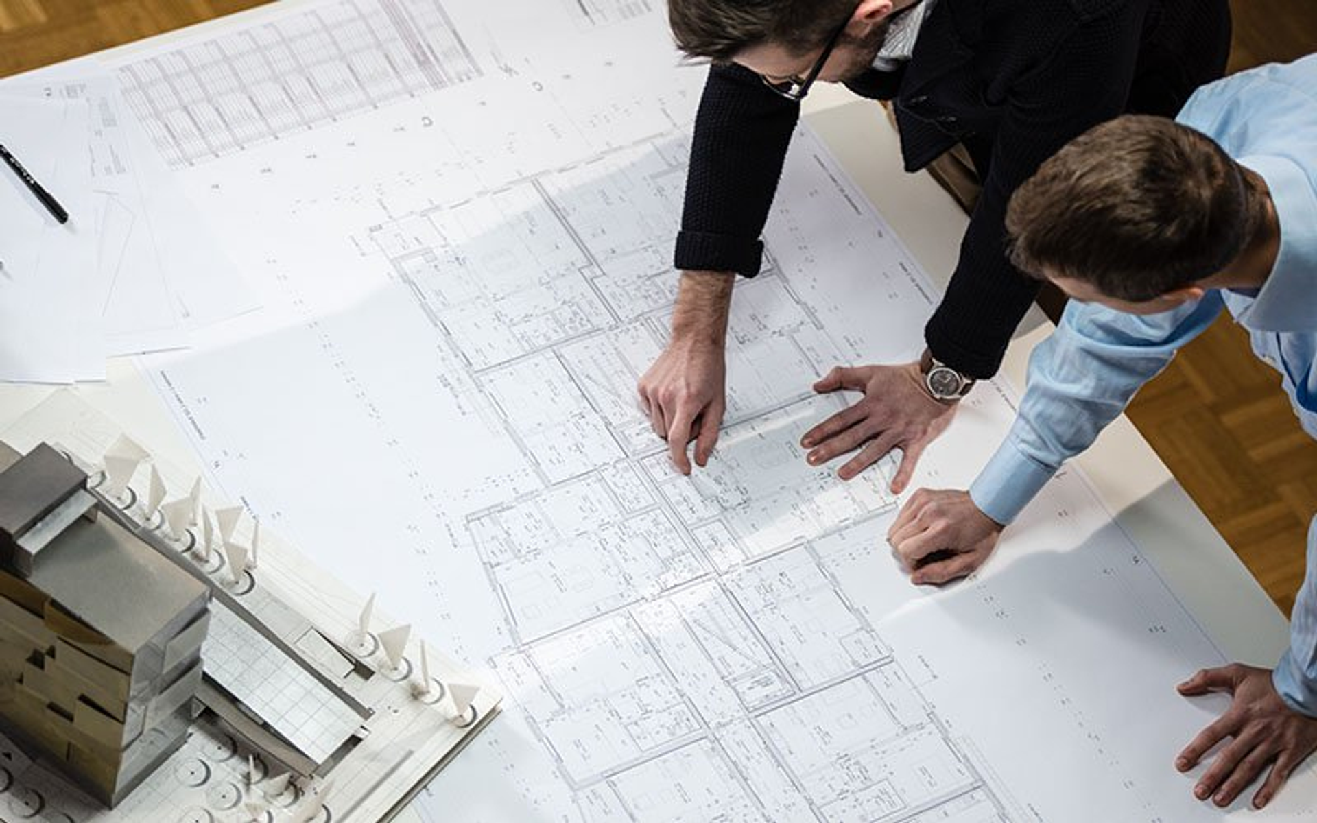 Zwei Männer betrachten Architekturpläne auf einem weißen Tisch. Gebäudemodell links.