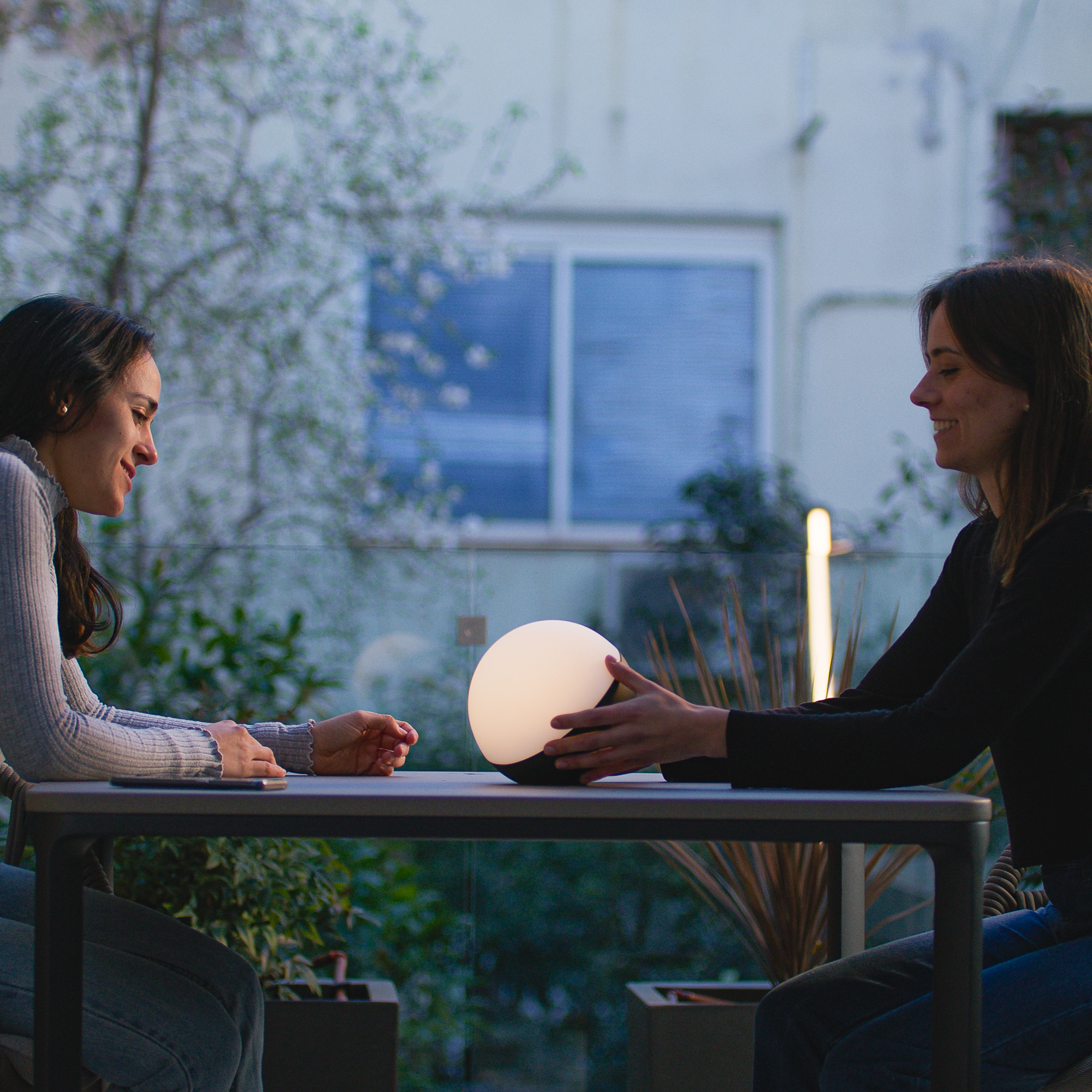 Dos mujeres en una mesa con una lámpara esférica encendida. Escena nocturna, fondo verde.