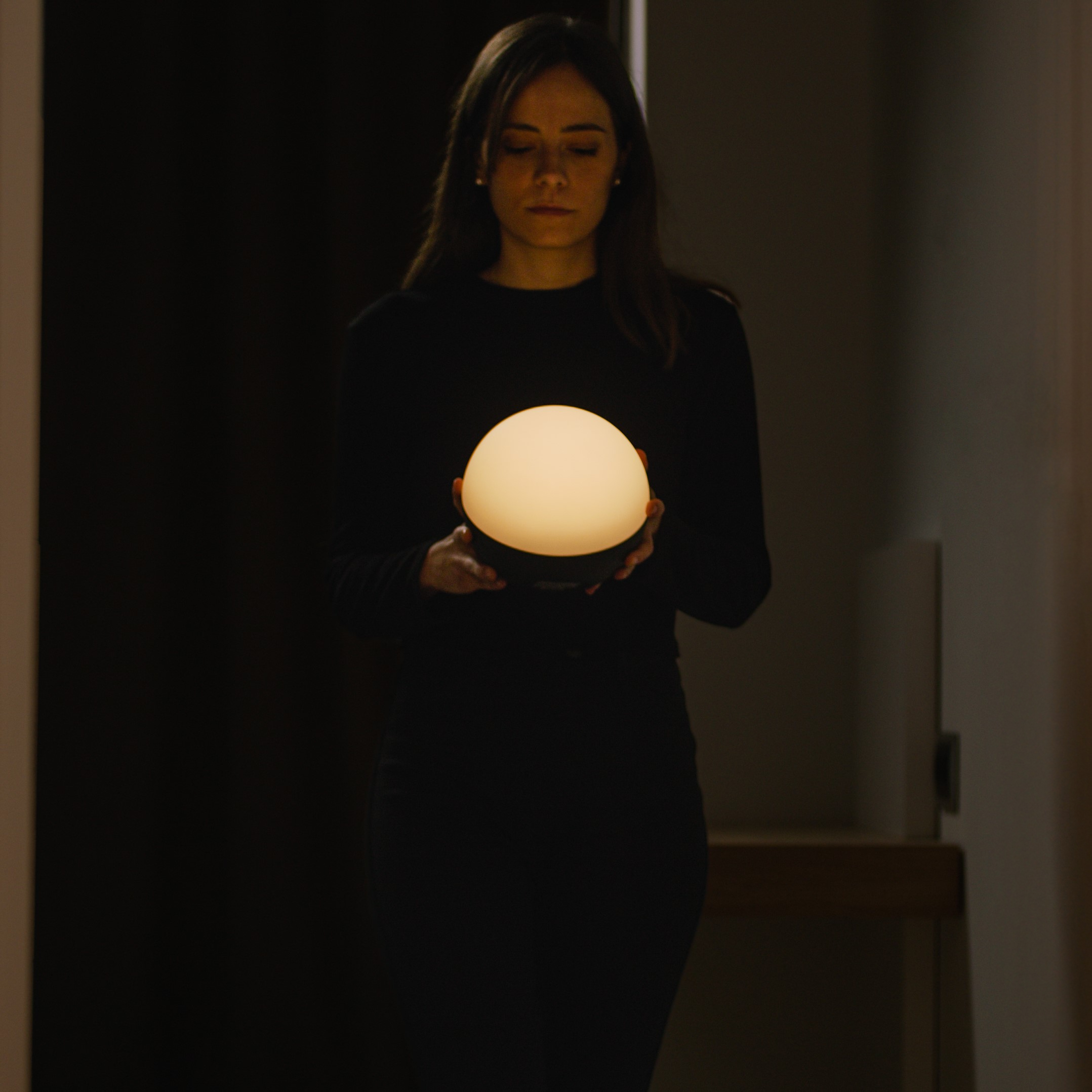 Una mujer con un vestido negro sostiene un orbe brillante en un pasillo oscuro.