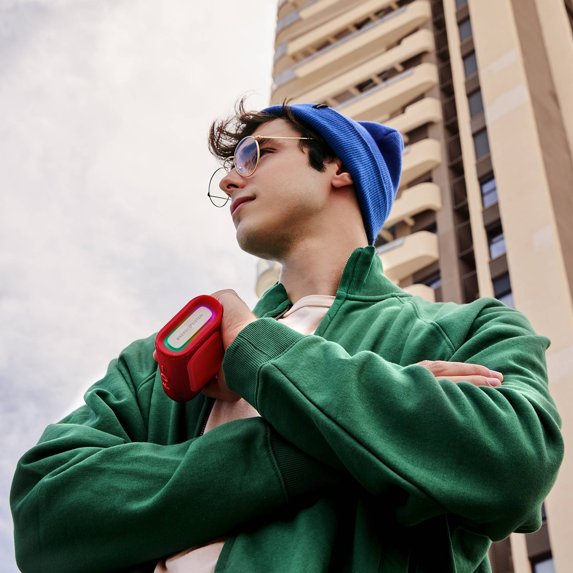 Un joven sosteniendo un altavoz rojo, con gafas y un gorro azul, con un edificio detrás de él.