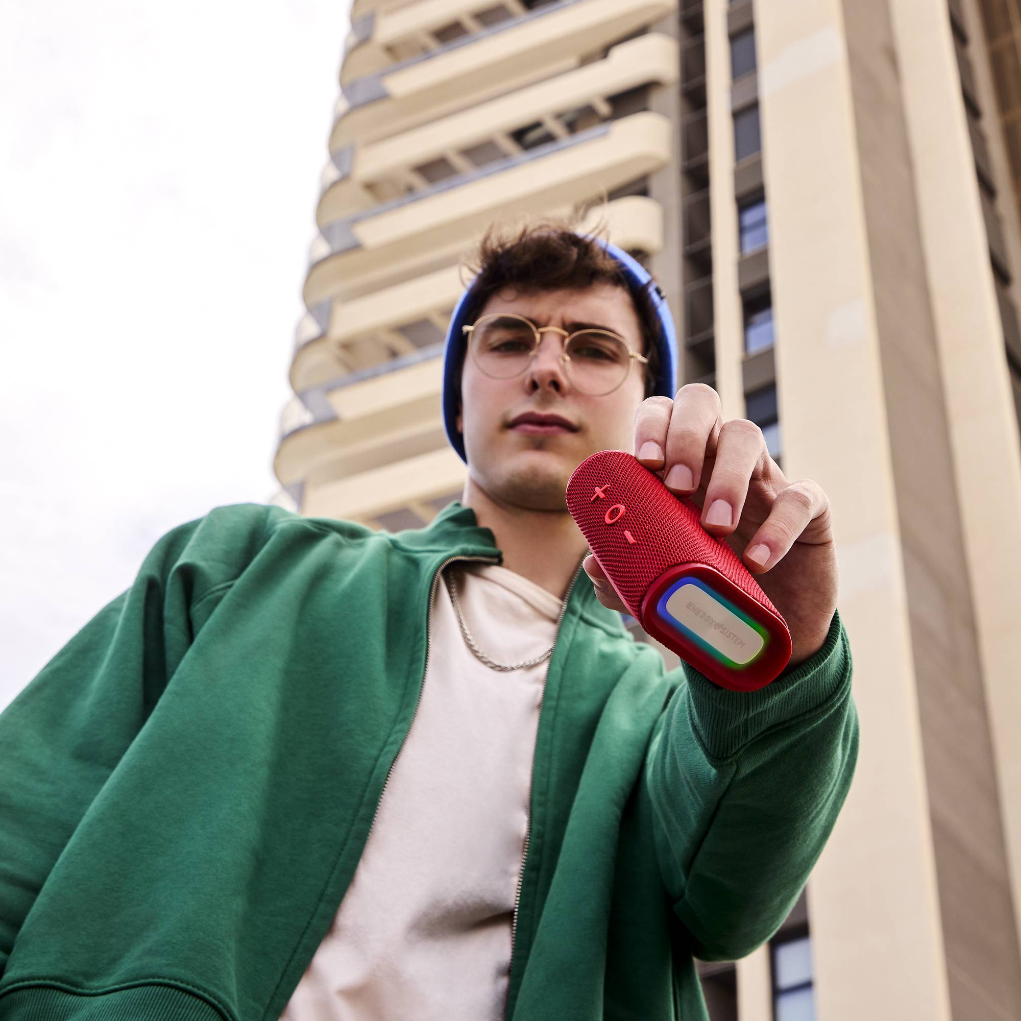 Un hombre sosteniendo un altavoz portátil rojo, chaqueta verde, fondo de edificio.