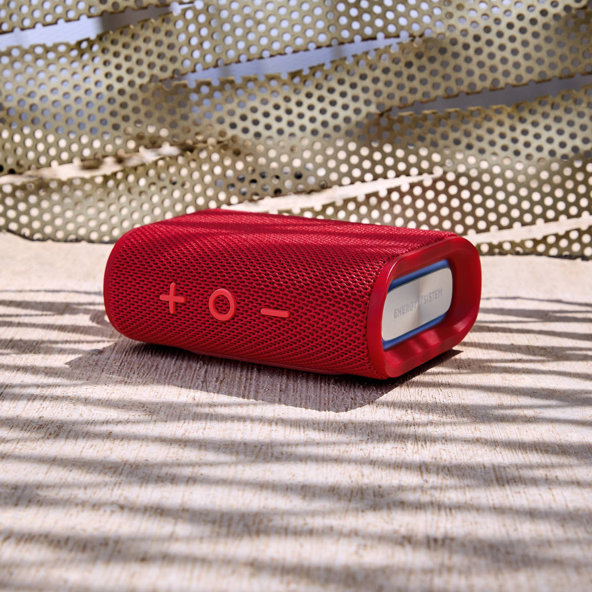 Un altavoz Bluetooth portátil rojo con botones, colocado sobre una superficie texturizada al aire libre.