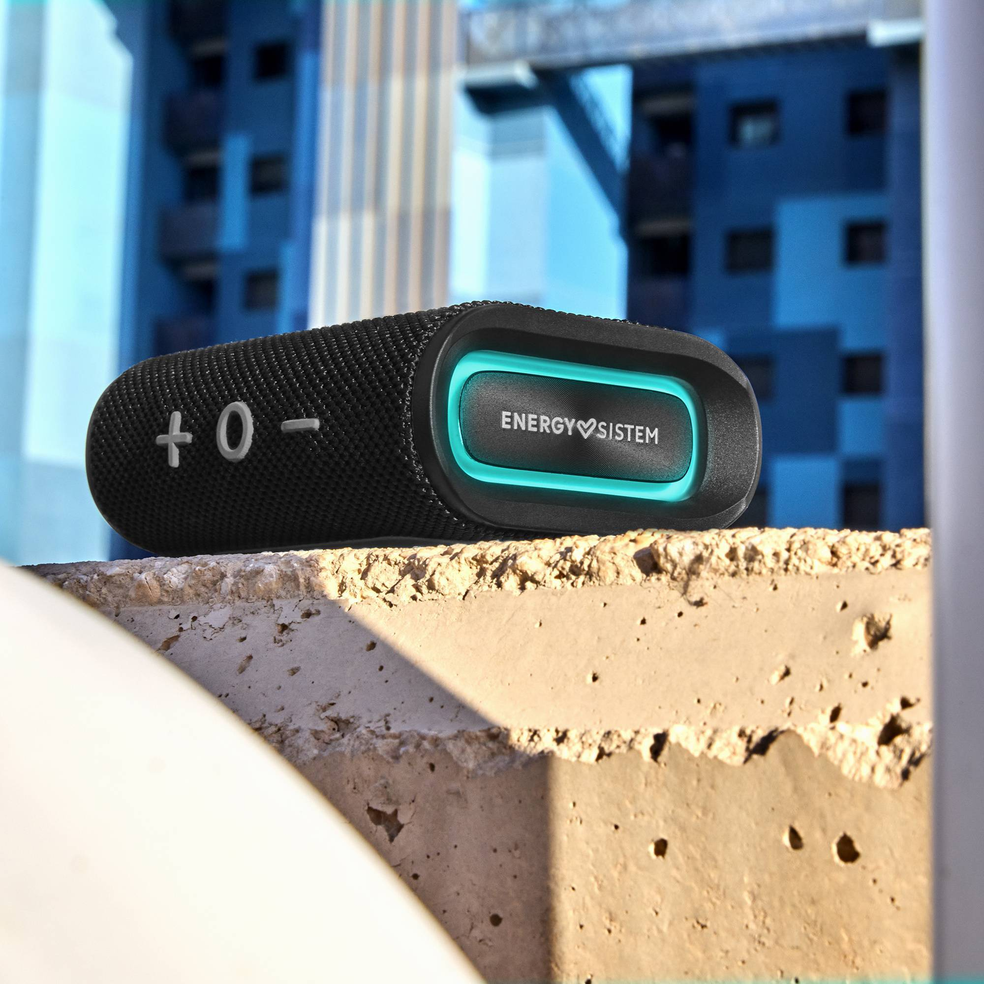 Altavoz negro con controles, luz turquesa y logotipo de Energy Sistem, sobre superficie de hormigón, con edificios detrás.