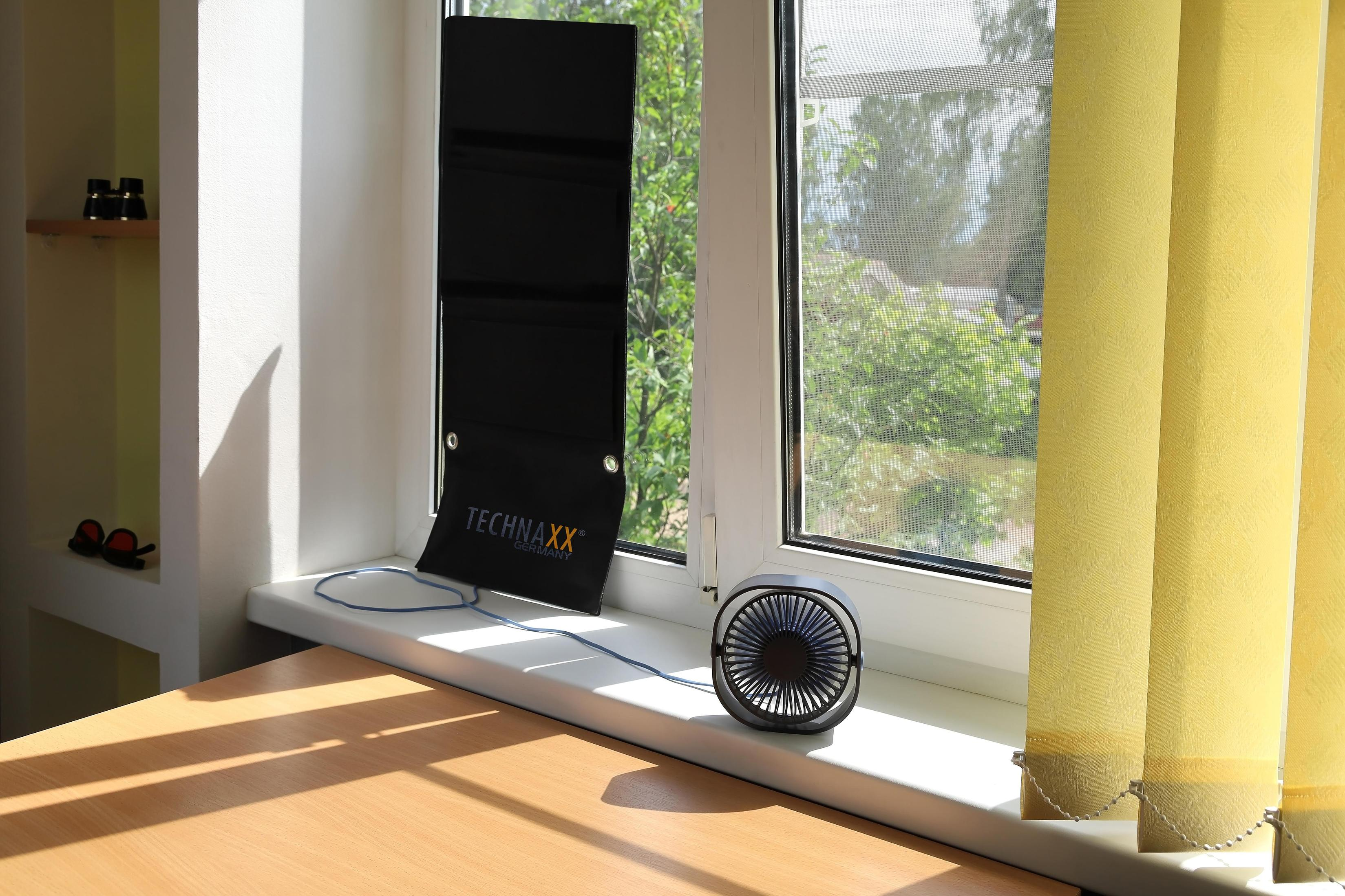 Okno z czarnym panelem słonecznym i małym wentylatorem na parapecie, ze stołem drewnianym na pierwszym planie.