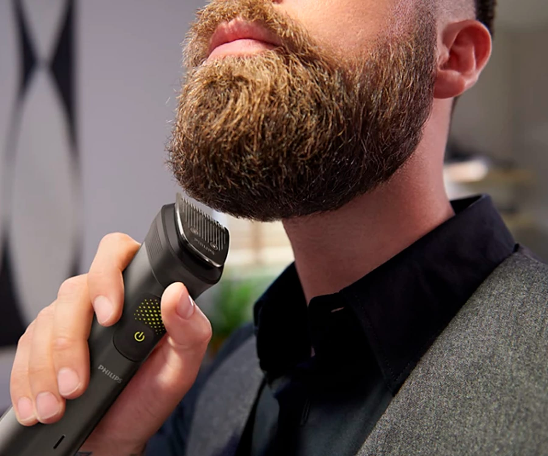 Mężczyzna przycina brodę trymerem elektrycznym. Zbliżenie twarzy. Szara koszula i rozmyte tło.