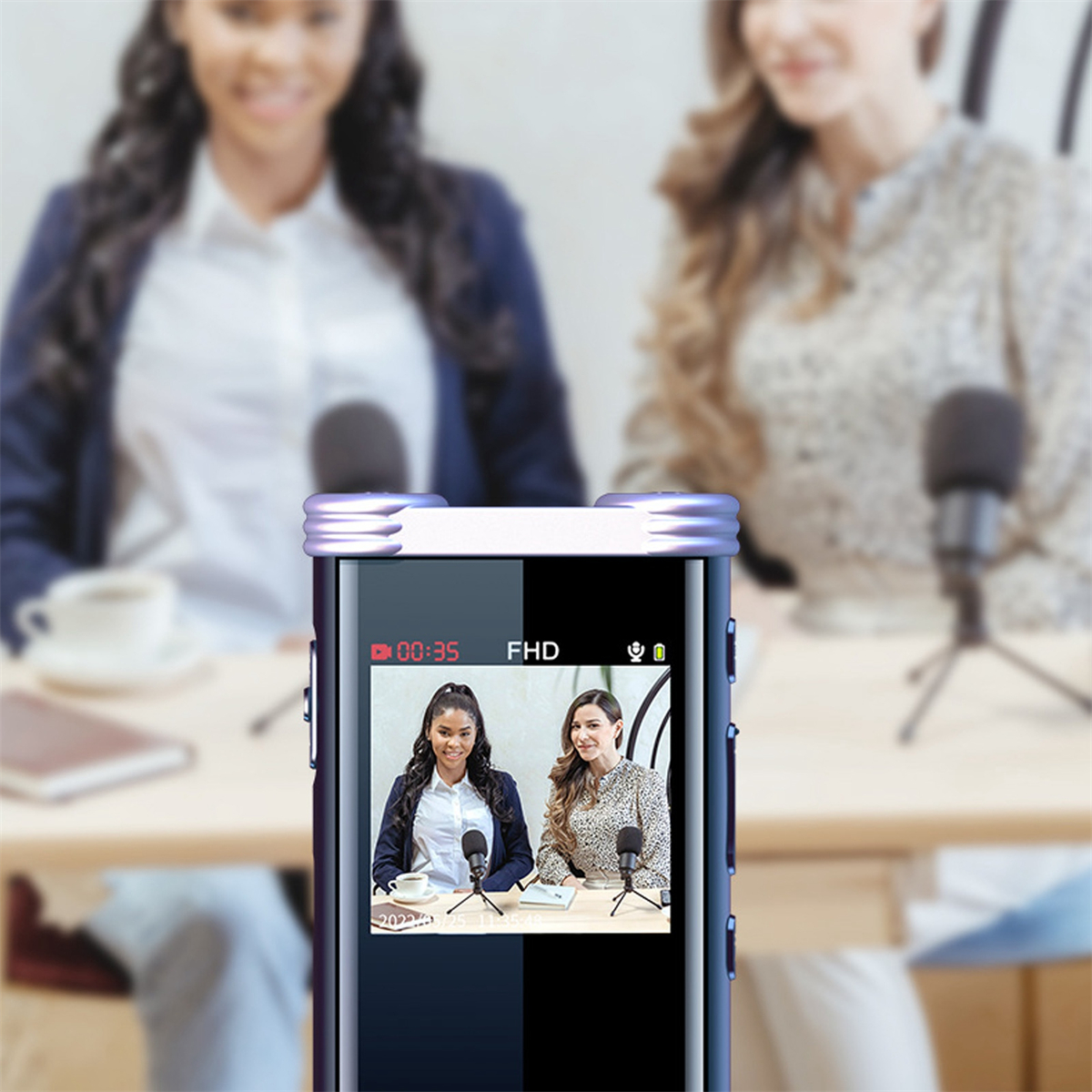 Zwei Frauen nehmen einen Podcast auf. Eine Frau ist auf dem Bildschirm eines Aufnahmegeräts zu sehen. Mikrofone und Tisch sind vorhanden.