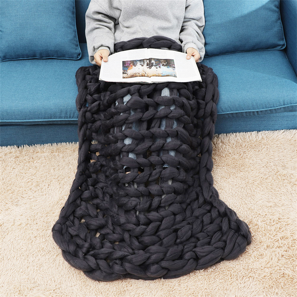 Person sitzt auf blauem Sofa, bedeckt mit grauer, grob gestrickter Decke, liest ein Buch. Beiger Teppich.