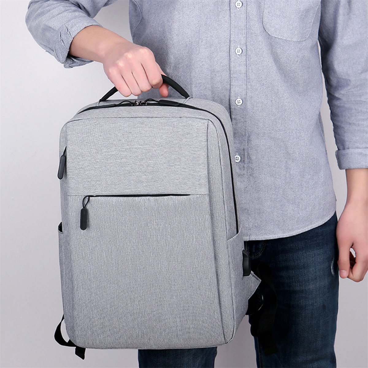 Mann hält einen grauen Rucksack. Der Rucksack hat eine Vordertasche, einen Griff oben und einen USB-Anschluss.