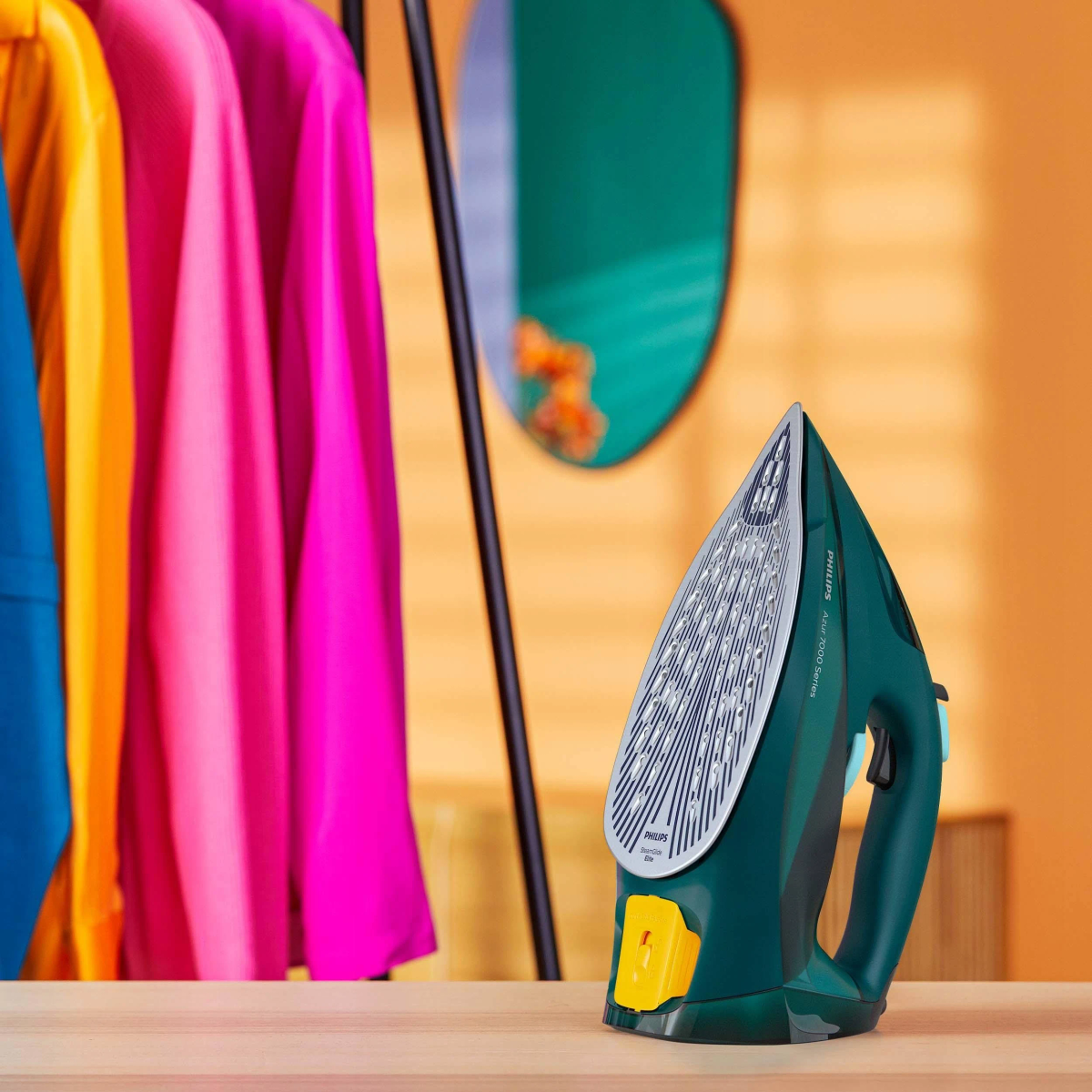 Zielone żelazko z żółtym guzikiem, wieszak na ubrania i lustro na pomarańczowej ścianie.