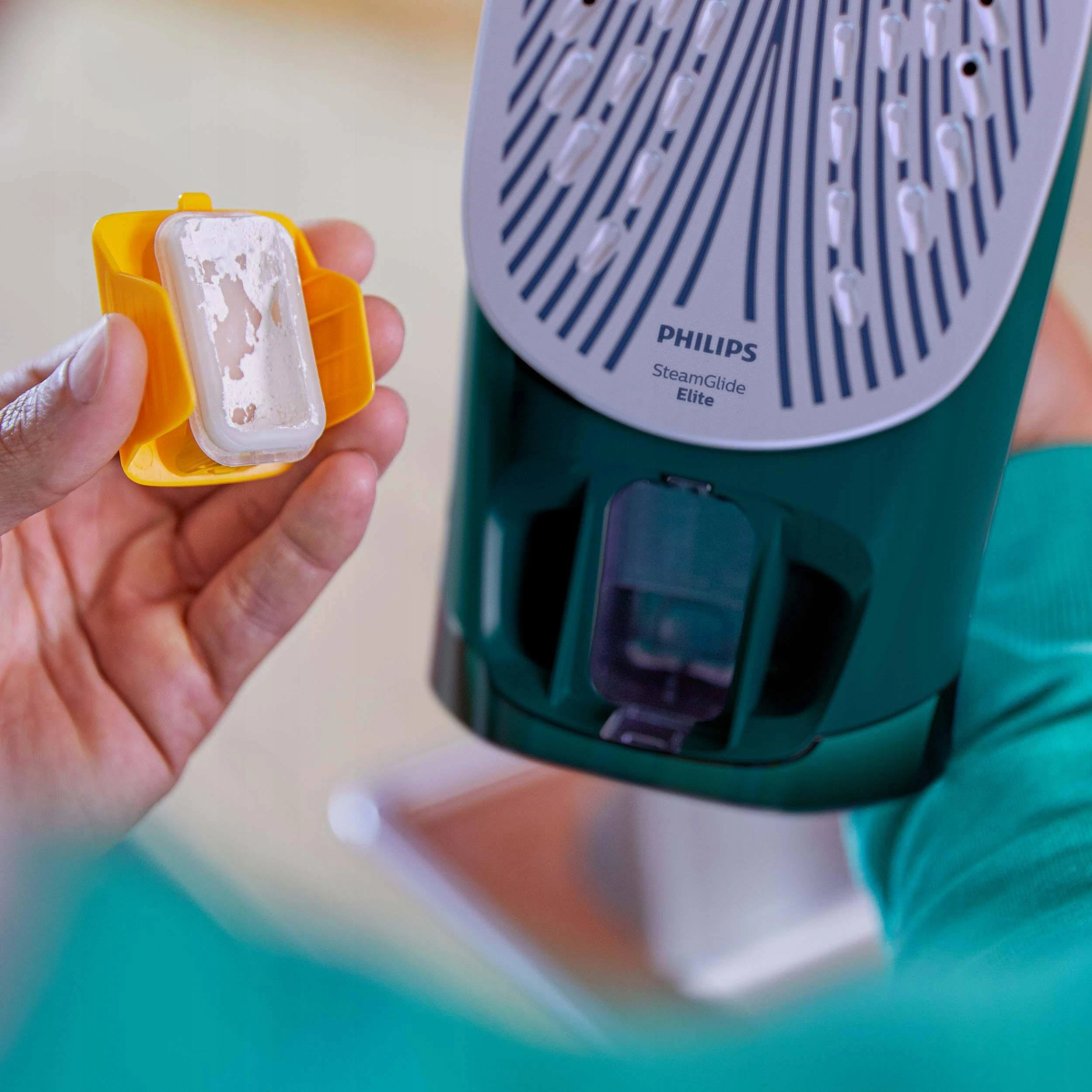 Ręka trzyma żółty pojemnik z białym filtrem. Zielone i białe żelazko w tle.